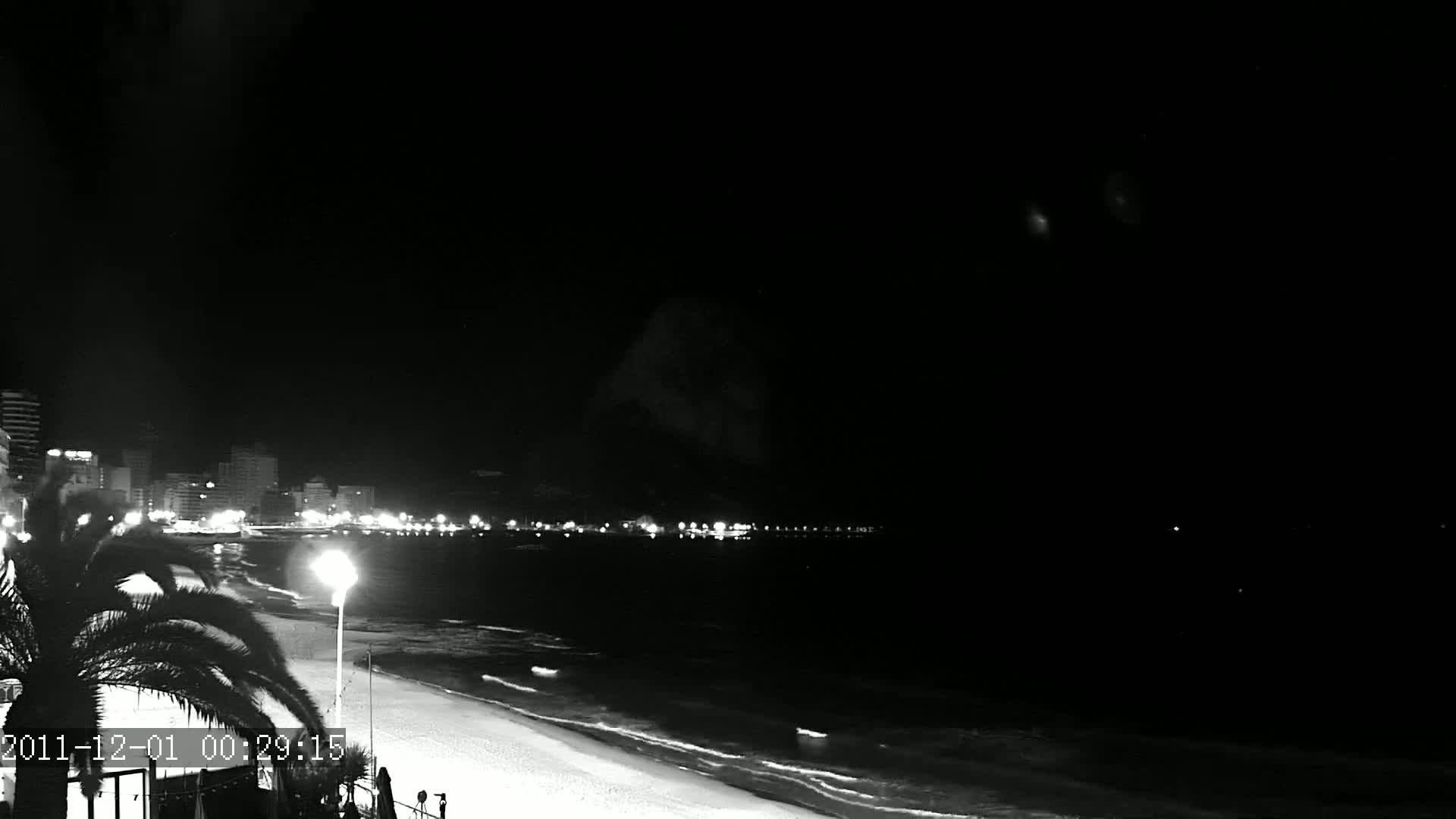 A nighttime, black and white view of a city skyline across a calm ocean with a sandy beach in the foreground.
