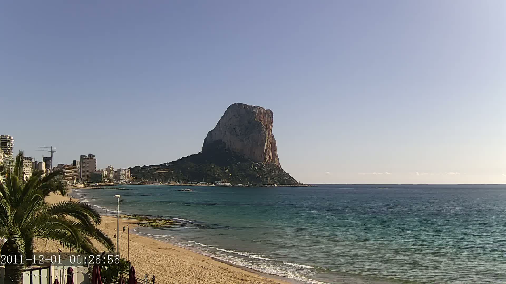 A large, imposing rock formation is visible across a calm, teal-colored ocean from a sandy beach with a few people visible, under a clear blue sky.
