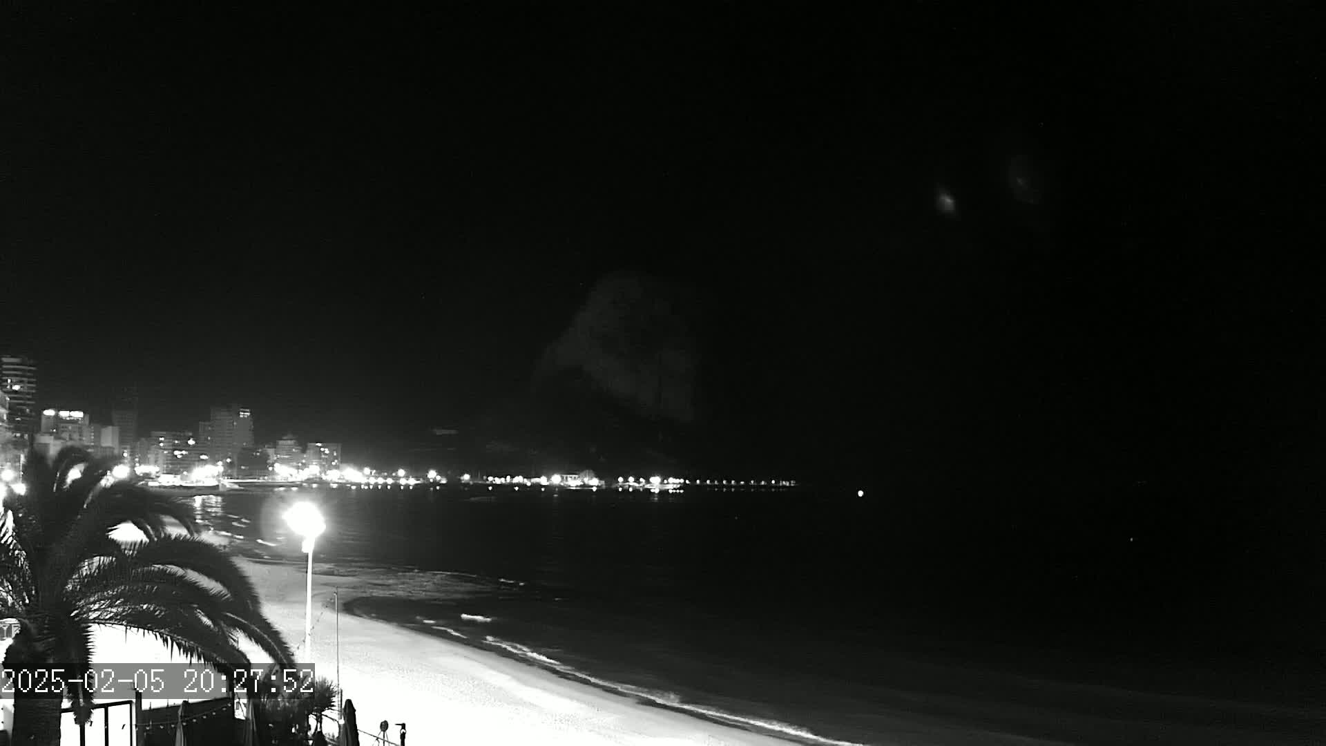 A nighttime beach scene shows a dark sky, lit city buildings across the water, and a sandy beach in the foreground.
