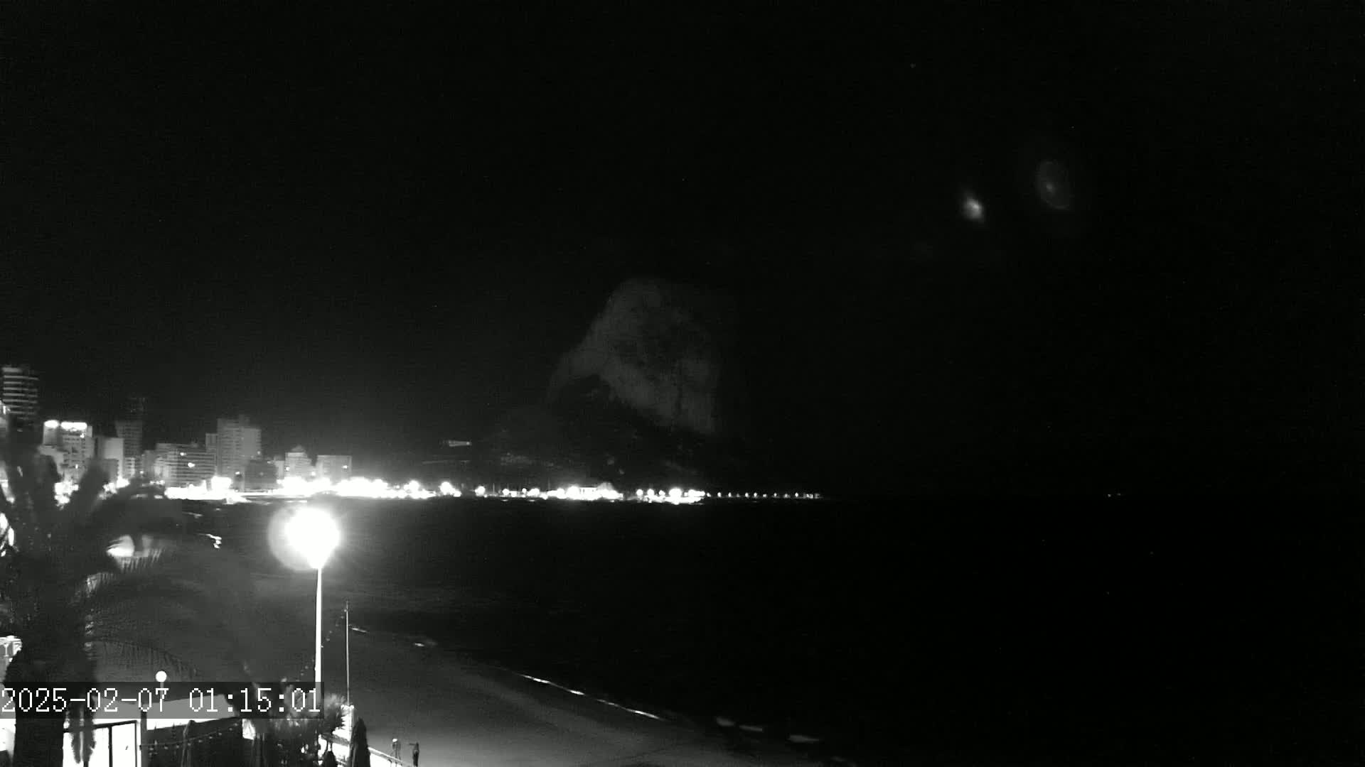 A nighttime, black and white view shows a dark ocean, a shoreline with lights from buildings, and a large dark hill or mountain in the background under a clear, dark sky.