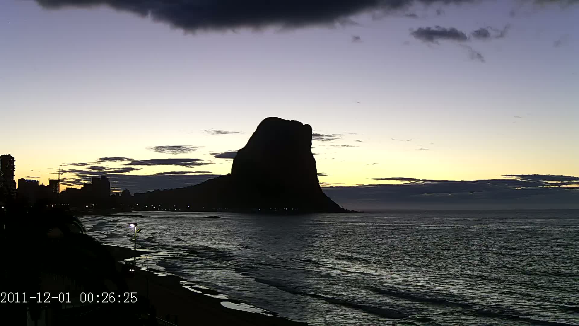 A large, dark silhouette of a rock formation stands prominently against a twilight sky over a dark ocean with small waves lapping a shoreline.