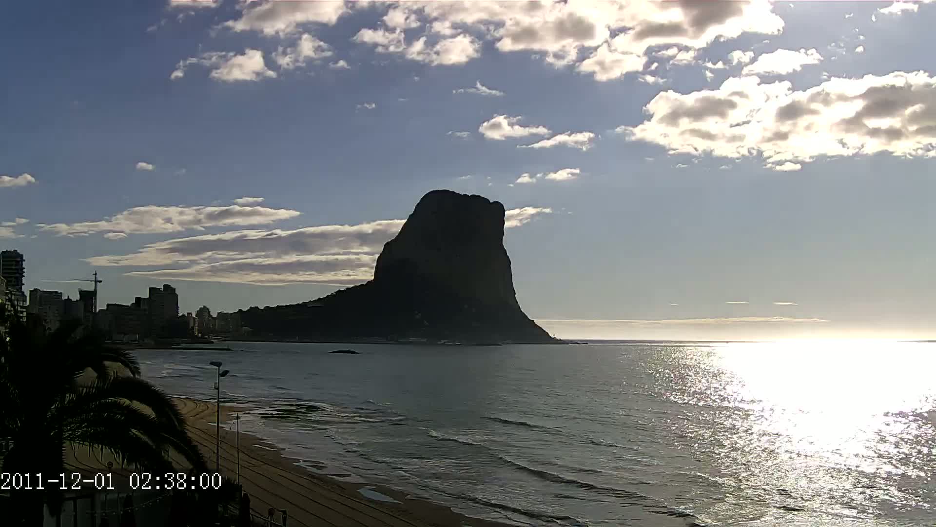 A large, dark mountain is silhouetted against a partly cloudy sky over a calm ocean with a sandy beach and a shoreline of buildings in the distance.