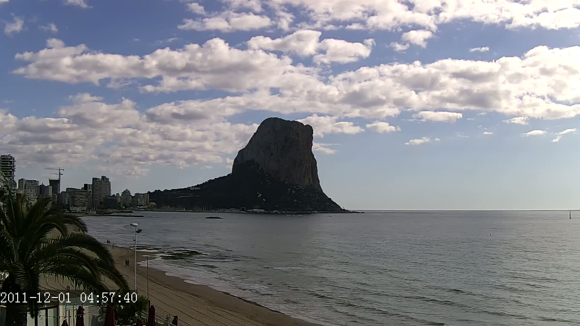 A large, dark rock formation rises from the sea under a partly cloudy sky, with a sandy beach and a cityscape visible in the foreground.