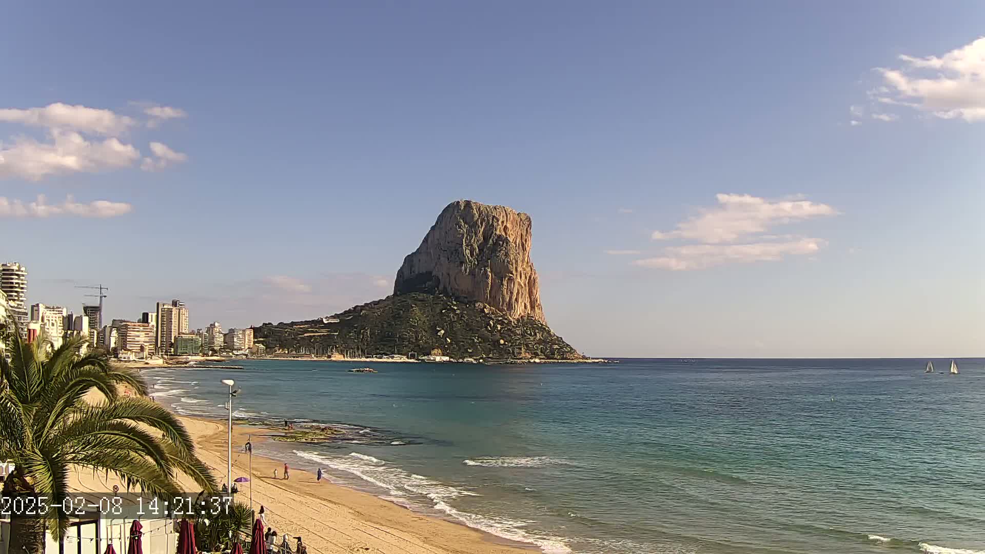 A sandy beach and calm ocean stretch out before a large, imposing rock formation, with a city skyline visible in the background under a partly cloudy sky.