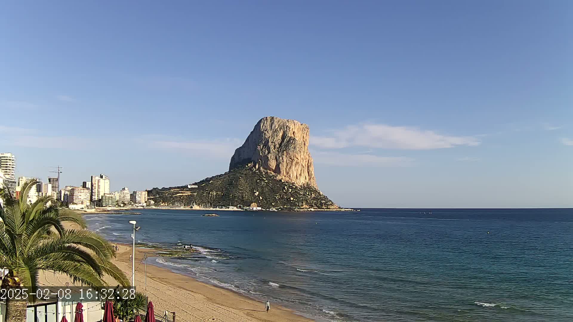 A large, rocky outcrop rises from the sea near a sandy beach and a line of buildings under a mostly clear blue sky.