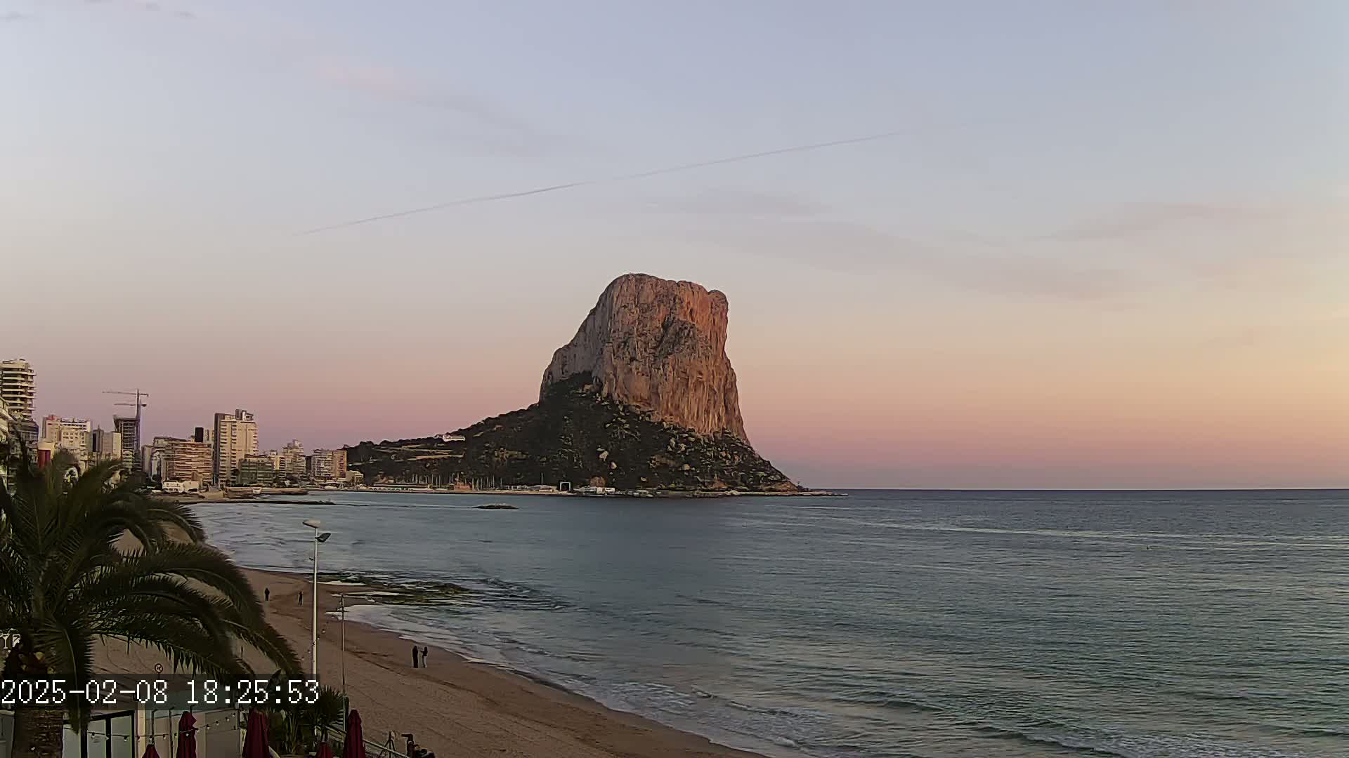 A large, rocky mountain rises from the sea at sunset, overlooking a beach with people and a city skyline in the distance under a clear, pale pink sky.