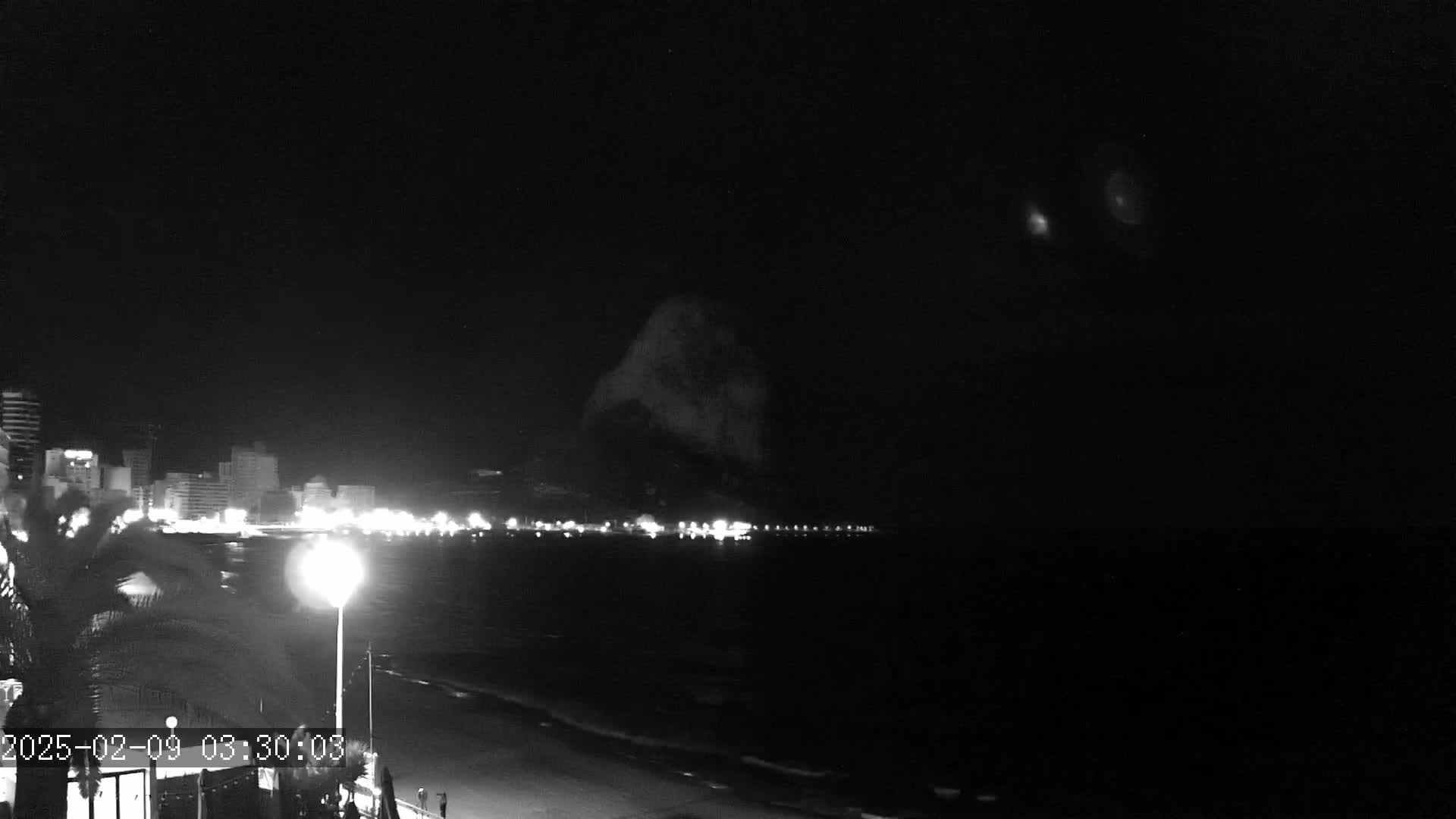 A nighttime, black-and-white view of a coastline with city lights across a dark body of water and a large, indistinct shape looming in the background.