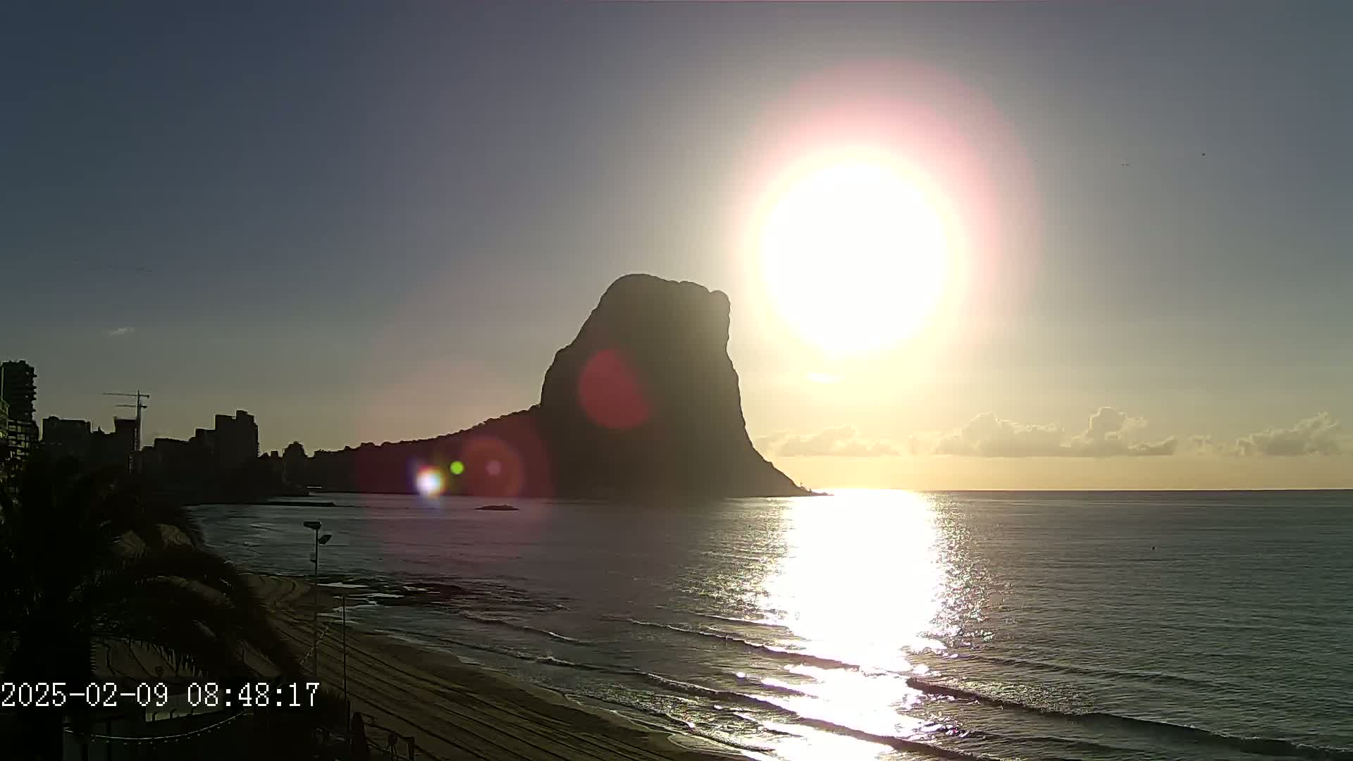 A large, dark rock formation rises from a calm sea at sunrise, silhouetted against a bright sun with a few clouds in the sky, a sandy beach and buildings visible in the distance.