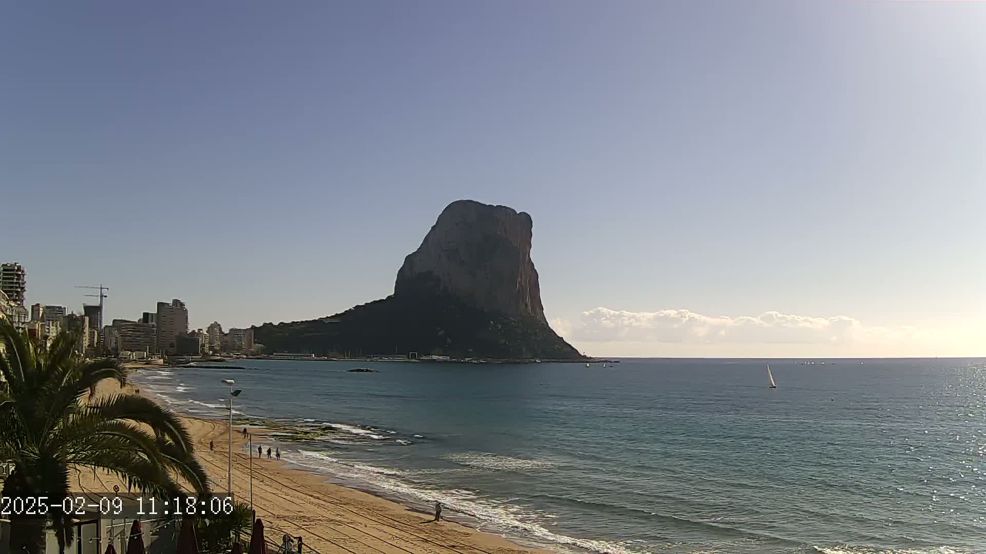 A large, dark rock formation rises from the calm, blue sea near a sandy beach and a line of buildings under a mostly clear sky with some clouds.