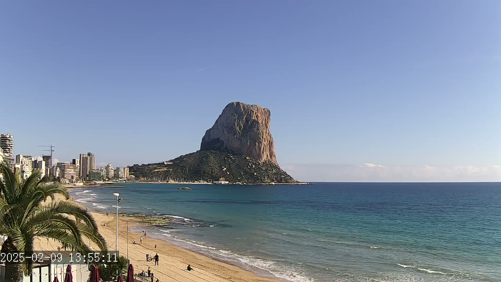 A large, rocky island rises from the calm, turquoise ocean, with a sandy beach and a city skyline visible in the background under a clear blue sky.