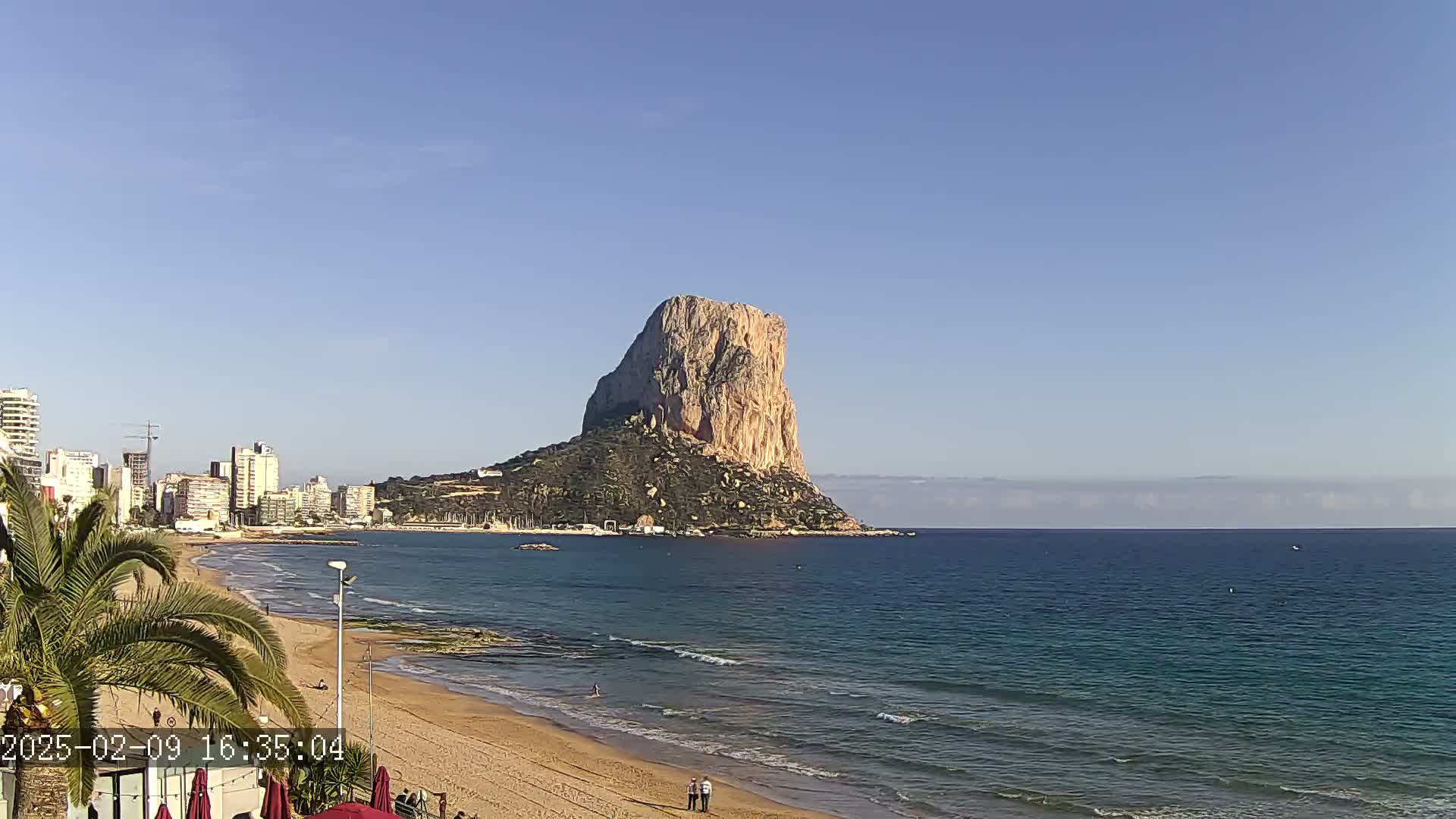 A large, light-brown rock formation rises from the sea near a sandy beach and a line of buildings under a clear blue sky.