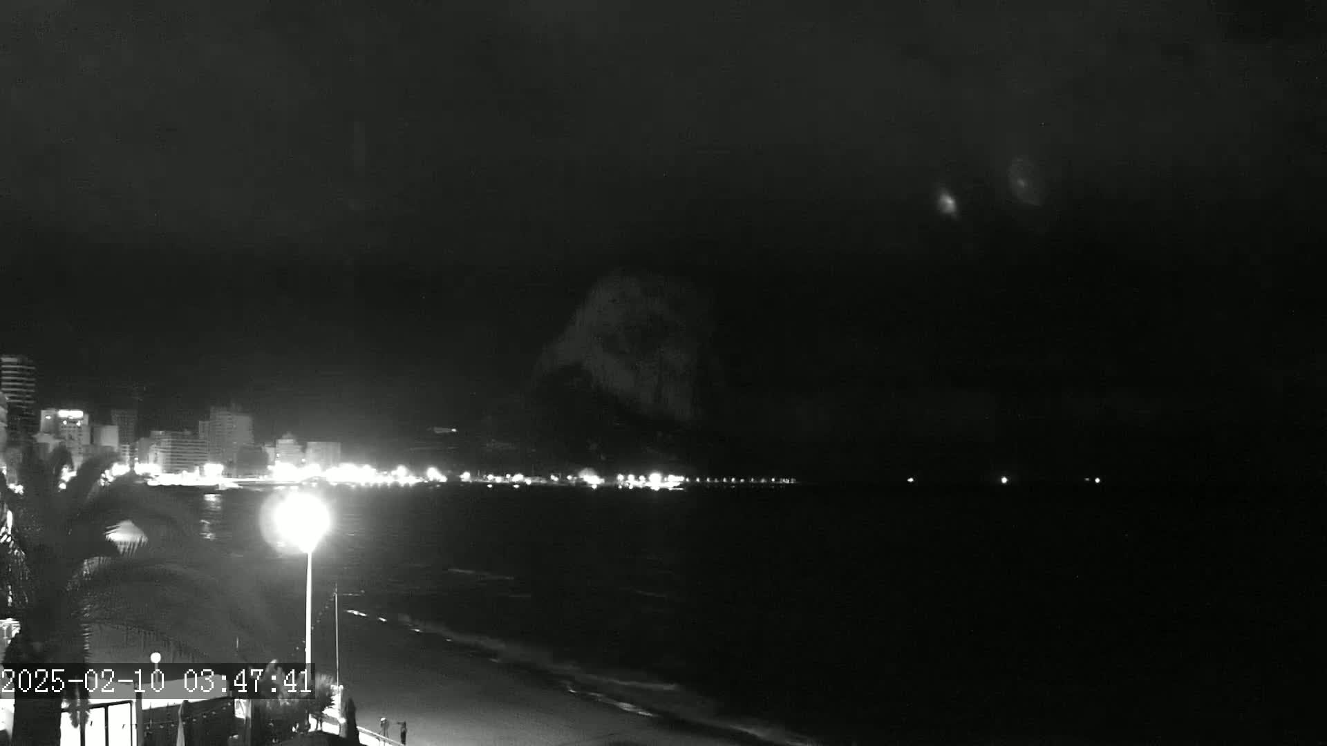 A nighttime, low-light view of a beach with a city skyline in the distance under a dark, cloudy sky.