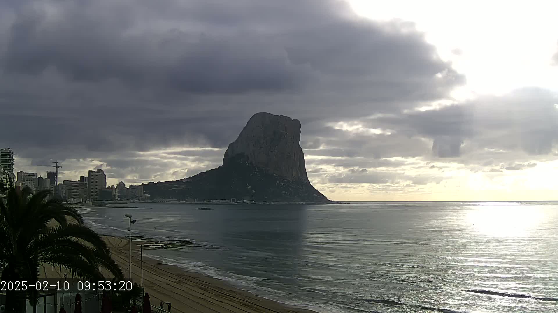 A large, dark rock formation rises from the sea under a cloudy sky, with a city skyline visible to its left and a sandy beach in the foreground.