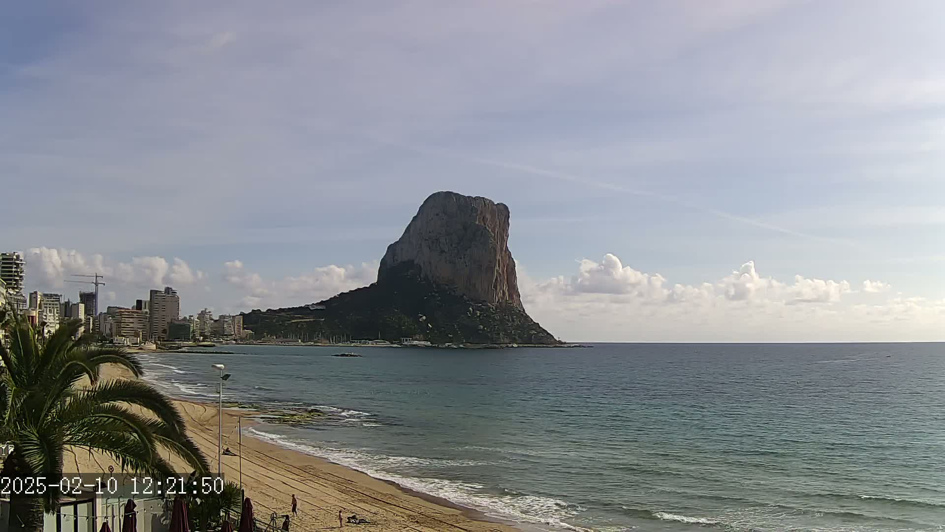A large, solitary rock formation rises from the sea near a sandy beach and a city skyline under a partly cloudy sky.