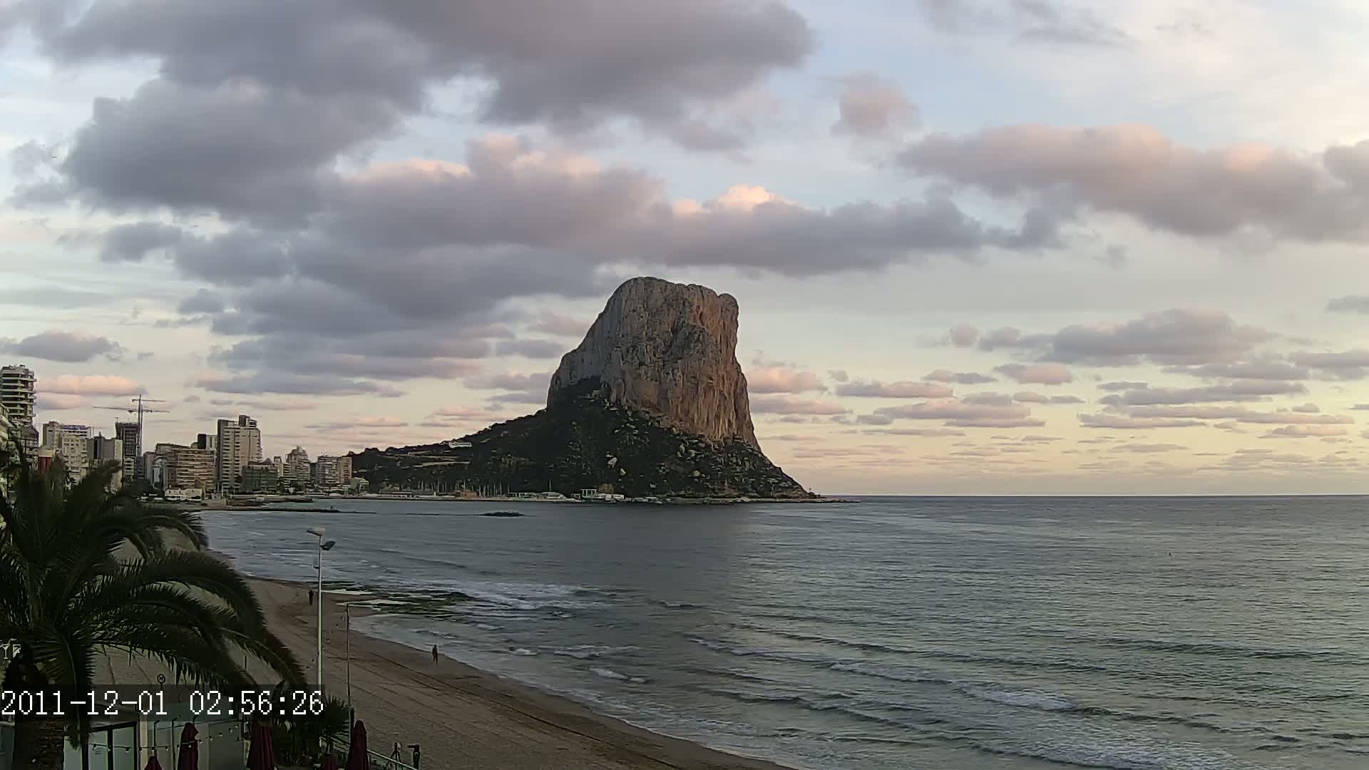 A large, rocky outcrop rises from the sea under a partly cloudy sky, with a city skyline and beach visible in the foreground.