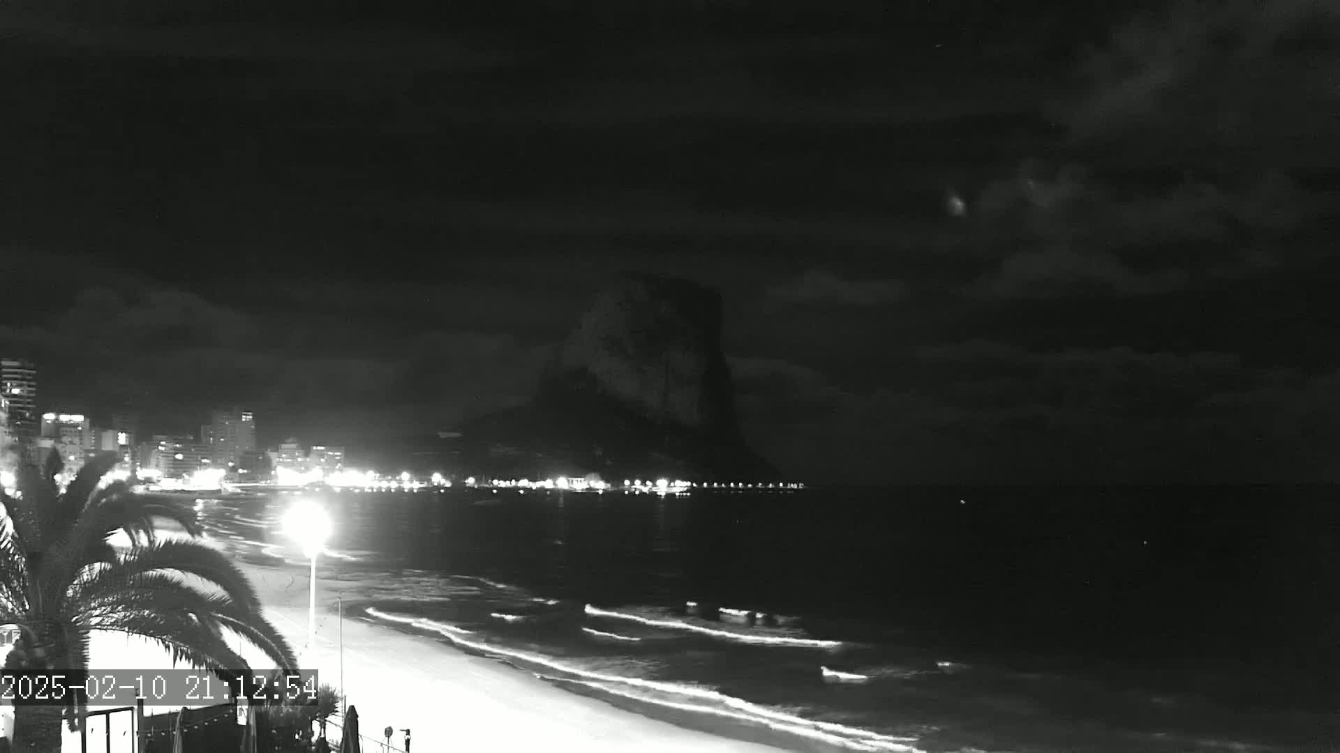 A nighttime beach scene shows a dark, cloudy sky over a city skyline and a large rock formation, with gentle waves lapping the shore.