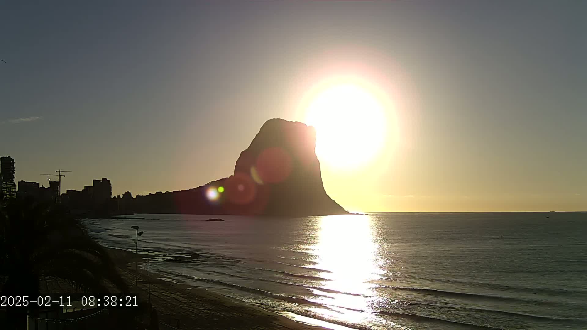 A large, dark rock formation silhouetted against a bright sunrise over a calm ocean with a beach and city skyline in the background.