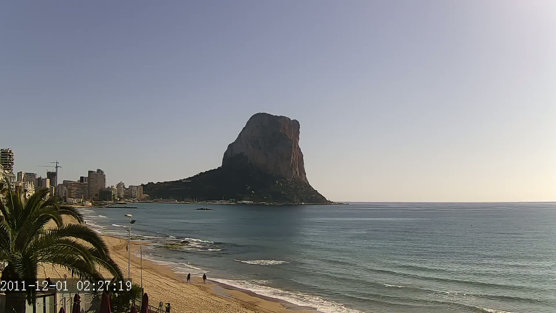 A large, dark rock formation rises from the ocean near a sandy beach with a few people walking, under a clear, light-blue sky.