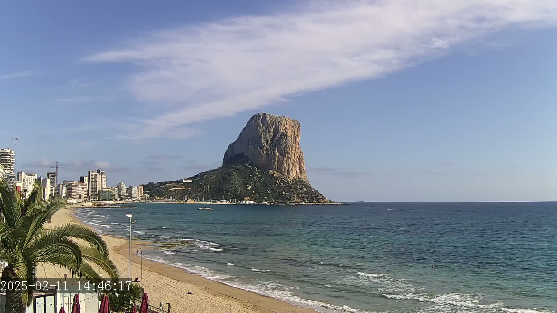 A large, rocky, tan-colored mountain rises from the ocean near a sandy beach with a cityscape in the background under a mostly clear, sunny sky.