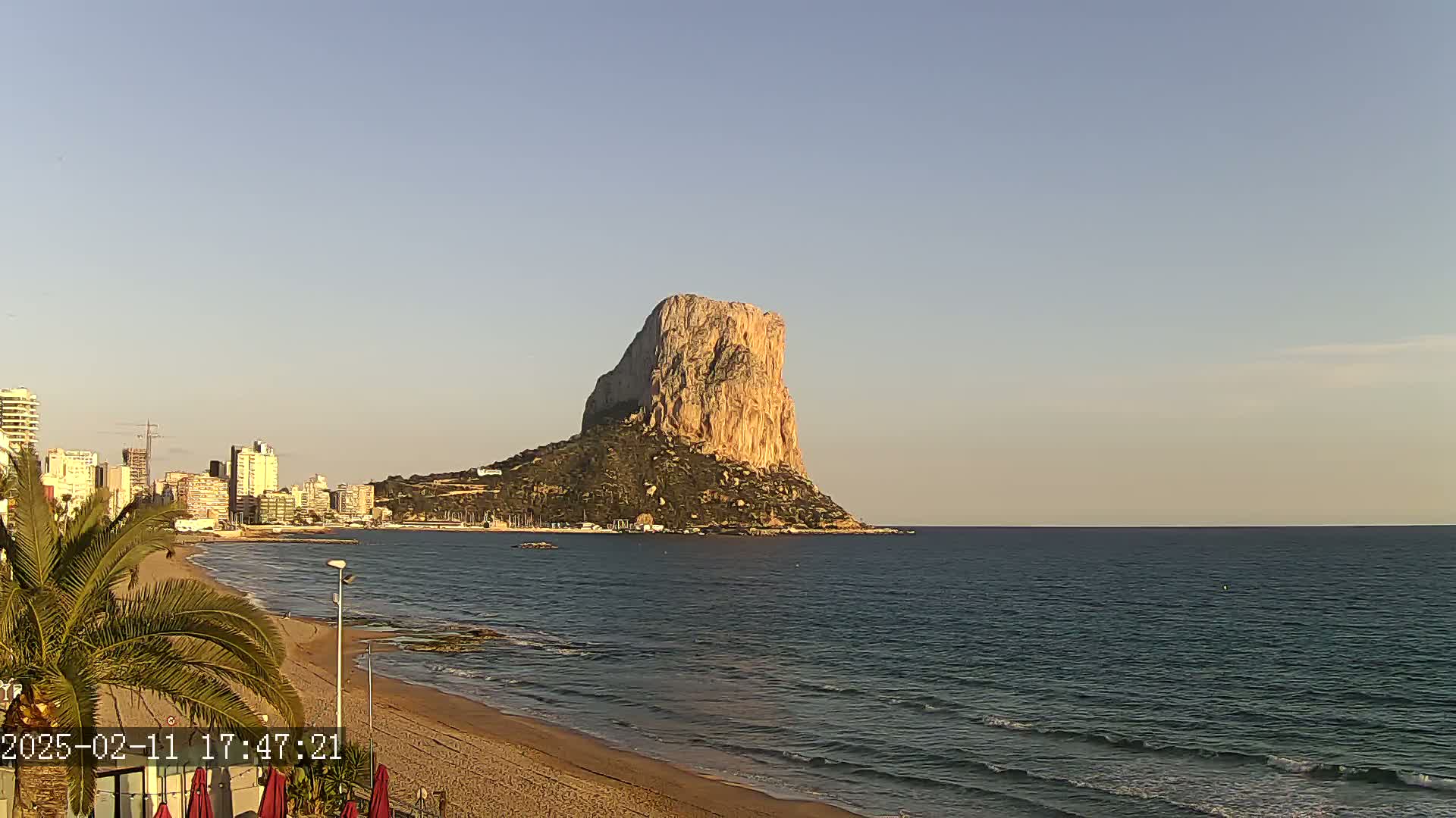 A large, tan rock formation rises from the sea near a sandy beach and a cityscape under a clear, light-blue sky.