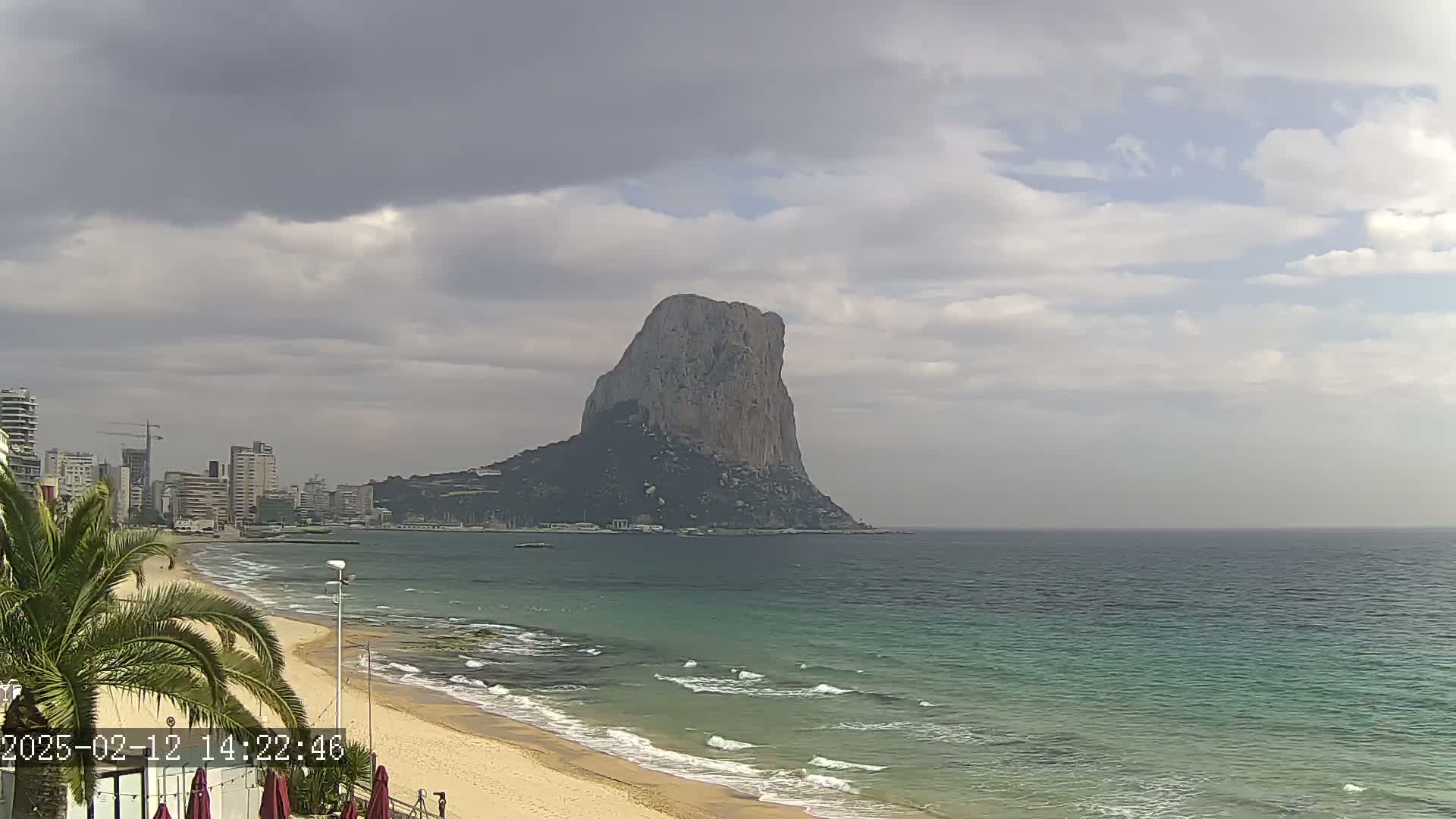 A sandy beach with gentle waves laps the shore, under a partly cloudy sky, with a large, imposing rock formation visible in the distance, and a cityscape in the background.