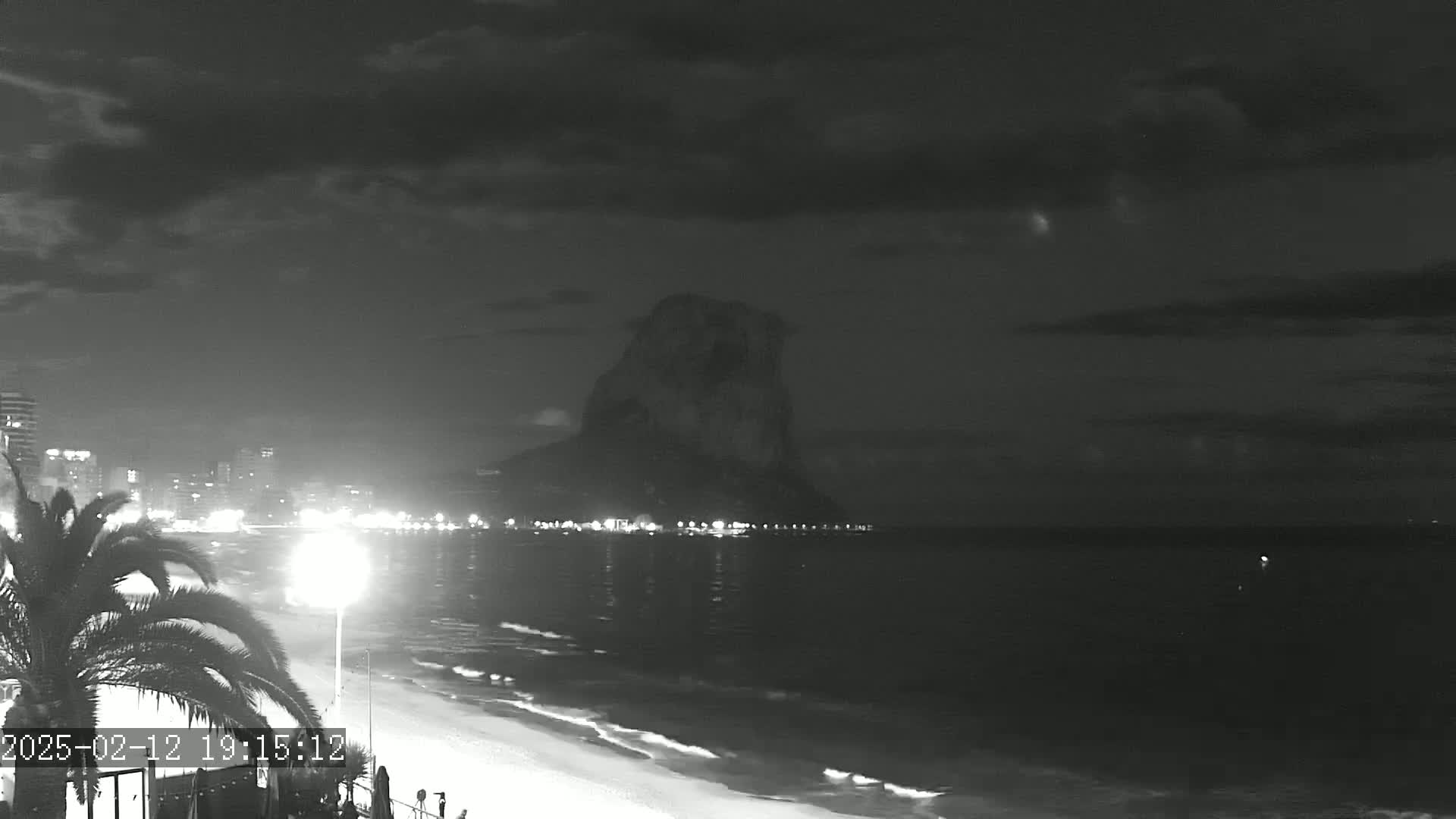 A nighttime beach scene shows a large dark rock formation across a calm body of water, with a city skyline and palm trees visible in the foreground under a cloudy sky.