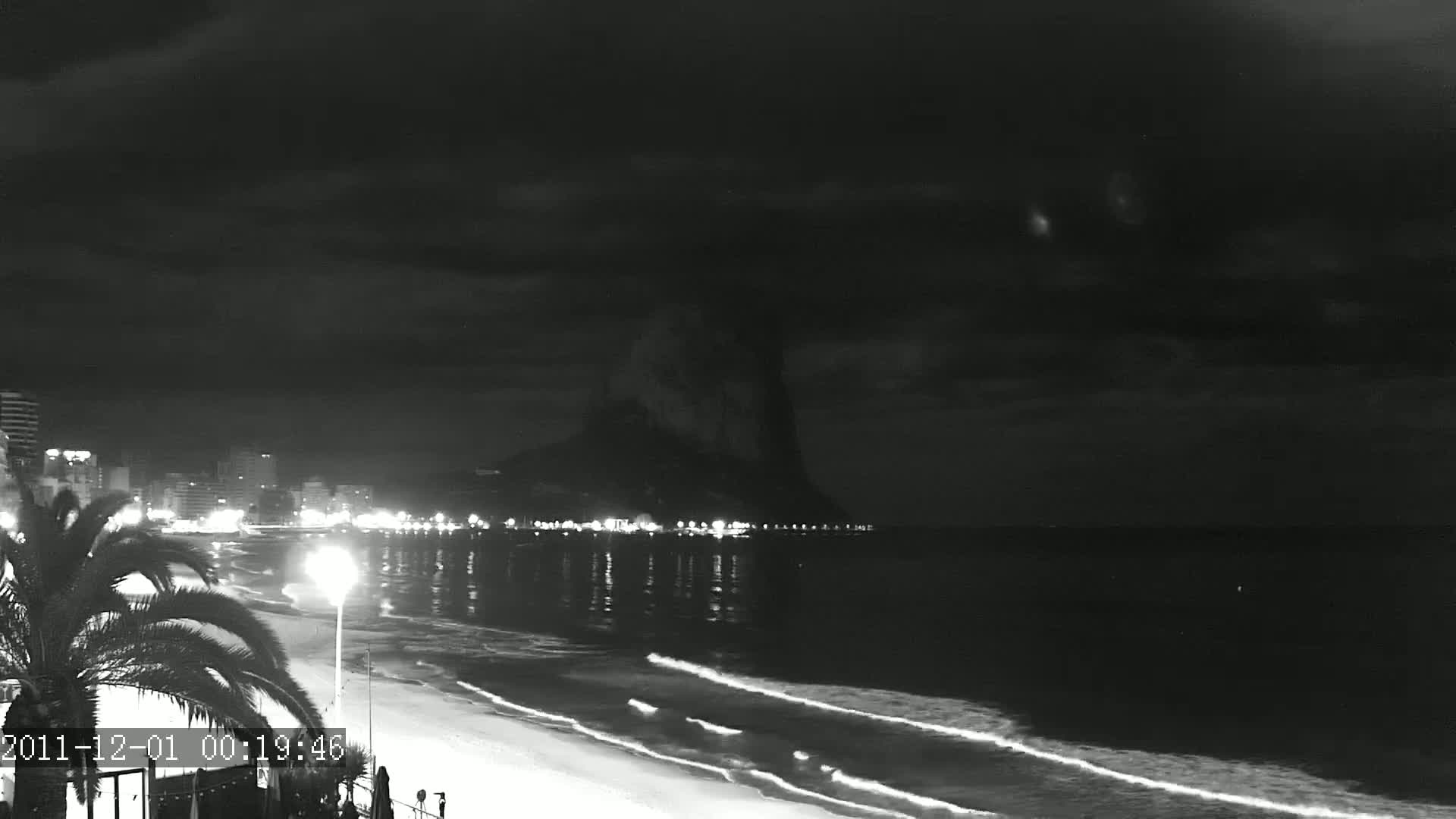 A nighttime beach scene shows a dark, cloudy sky over calm waves lapping a sandy shore, with a city skyline and a large, dark mountain in the background.