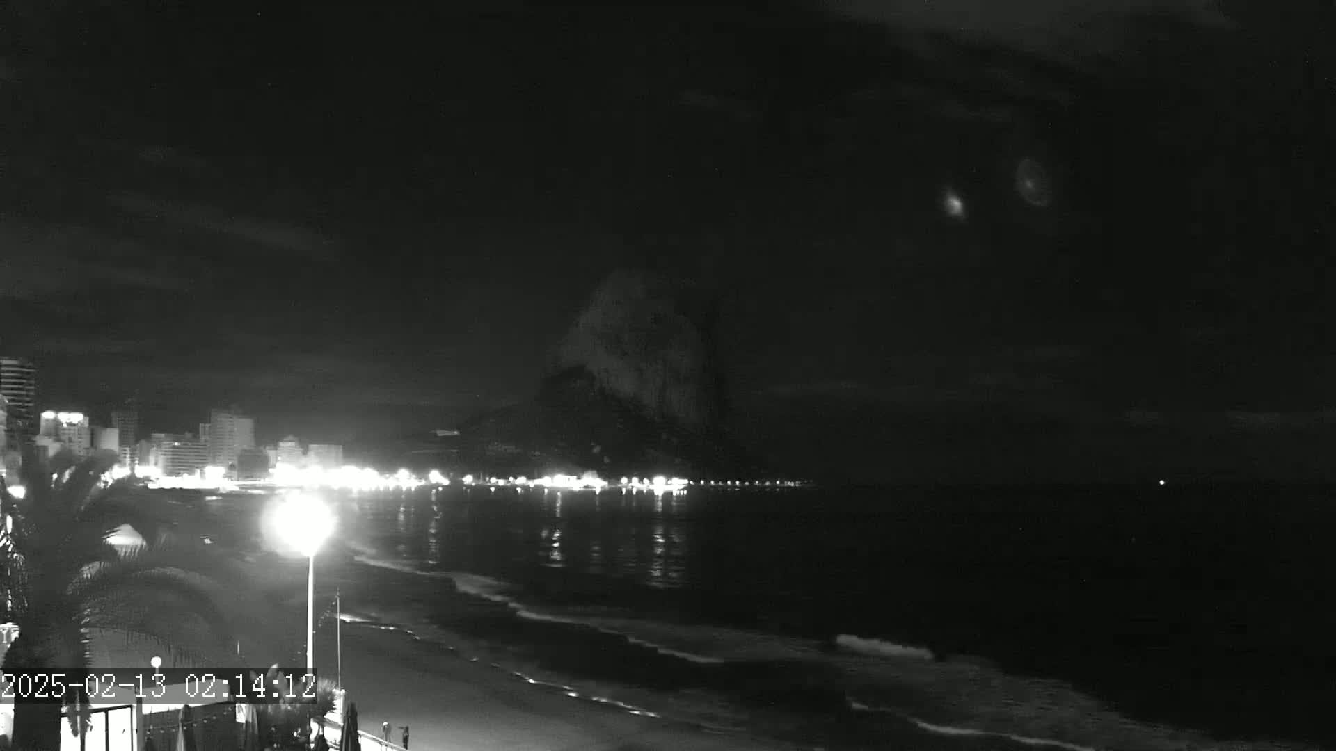 A nighttime beach scene shows a dark, cloudy sky above a dimly lit shoreline with buildings and a large, dark landmass in the distance, with gentle waves lapping the shore.