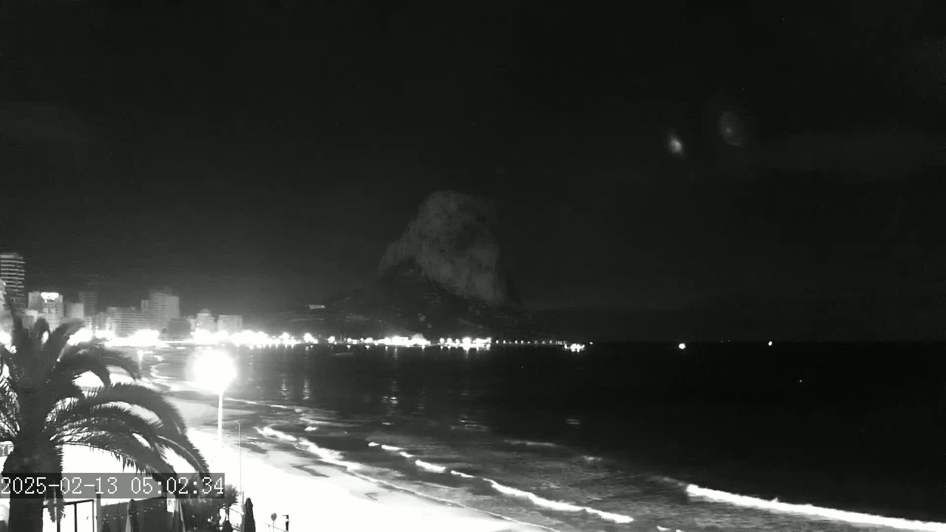 A nighttime beach scene shows a dark, rocky mountain in the distance, a shoreline with lights from buildings and streetlights, and gentle waves lapping on the sand under a clear, dark sky.