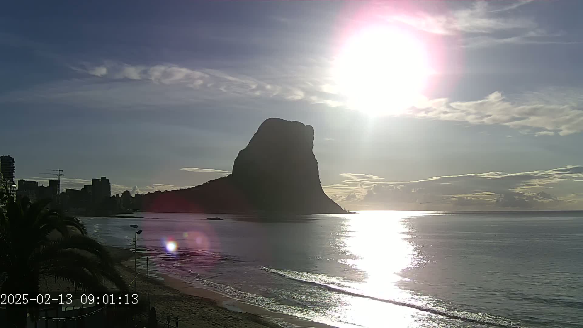 A sunrise illuminates a beach, city skyline, and a large, dark rock formation in the ocean under a partly cloudy sky.