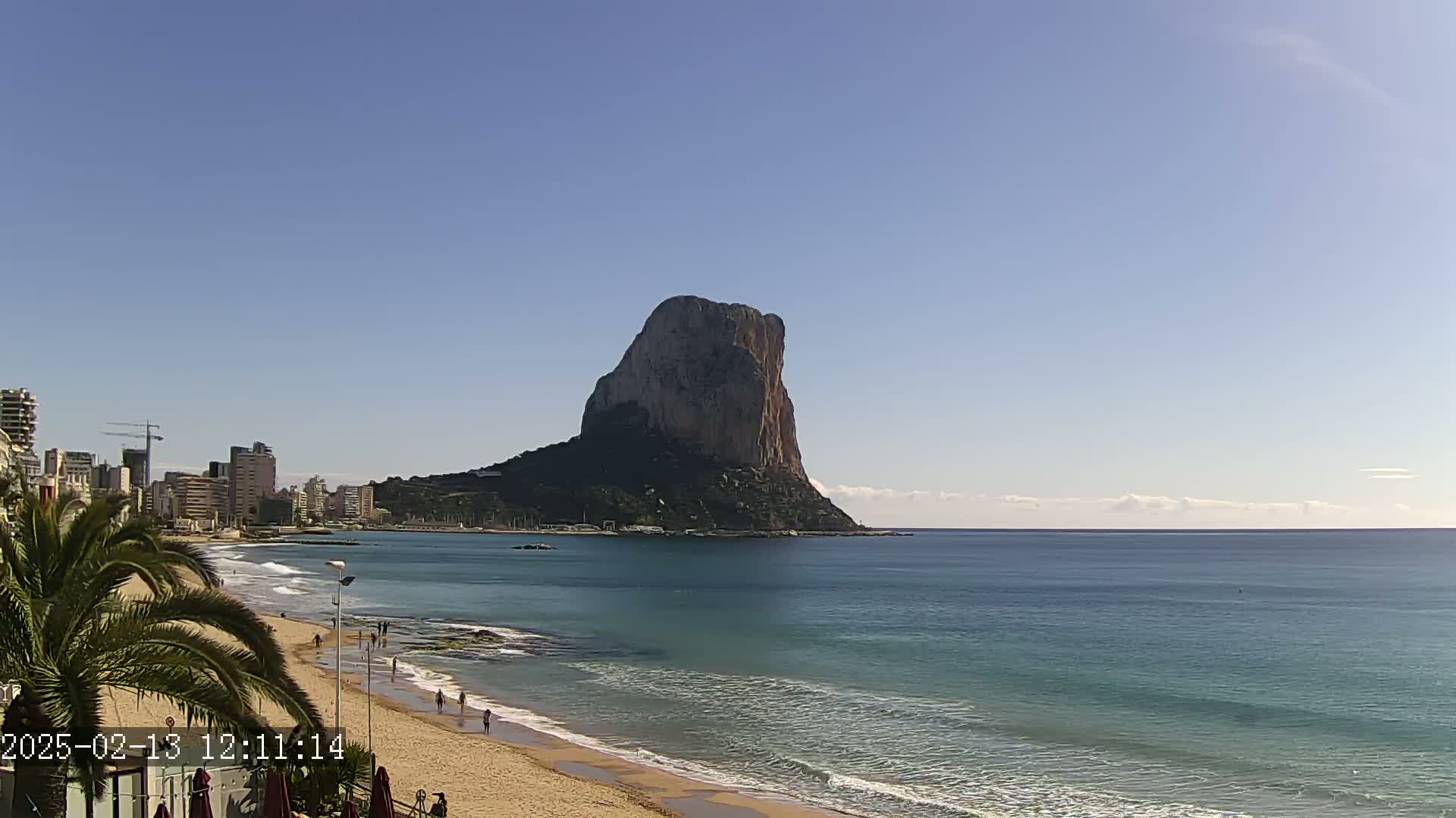 A large, rocky island rises from the calm, teal ocean next to a sandy beach and a city skyline under a clear blue sky.
