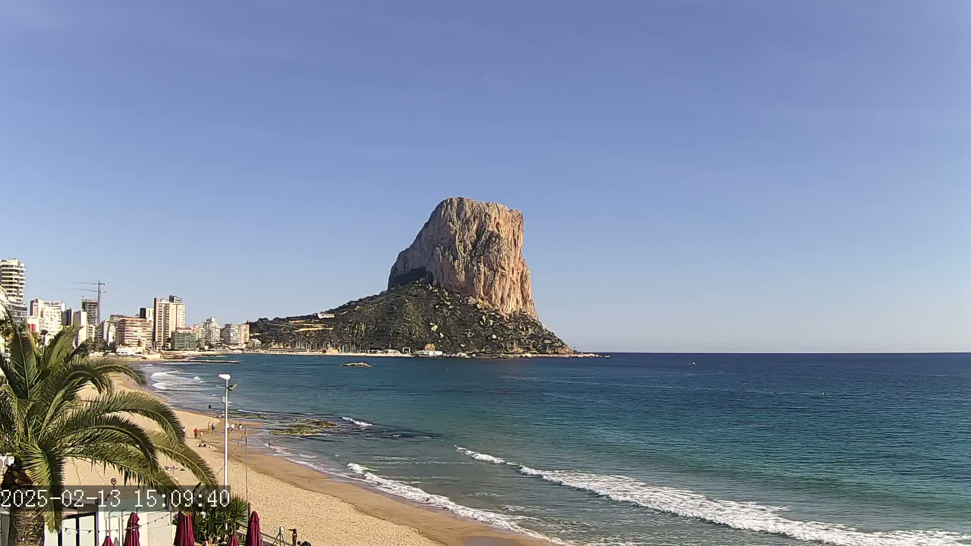 A large, rocky, tan-colored island sits in the ocean under a clear blue sky, with a sandy beach and buildings visible along the shore.