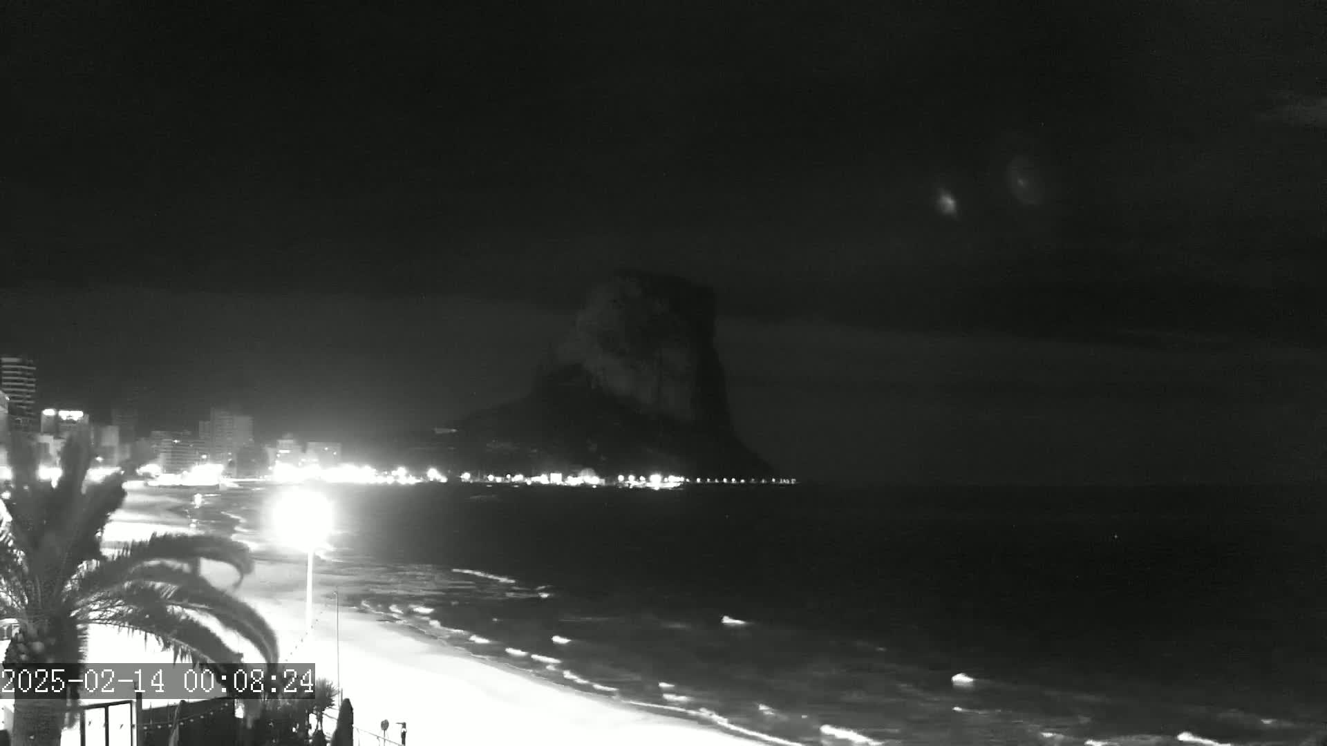 A nighttime, black-and-white view shows a beach with a city and a large, dark rock formation in the background, under a dark, cloudy sky.