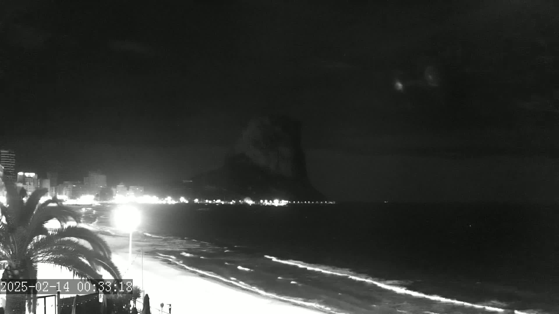 A nighttime, black-and-white view shows a dark, sandy beach with gentle waves lapping the shore under a mostly clear night sky, with a large, dark rock formation visible in the distance, and lights illuminating a city skyline along the beach.