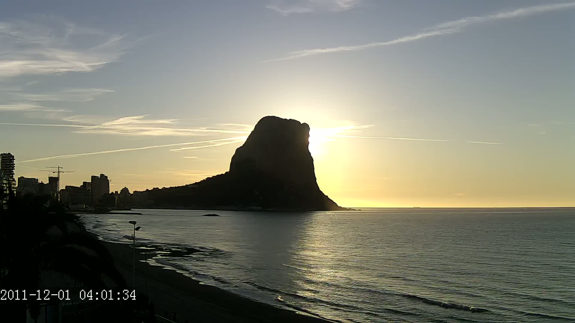 A large, dark rock formation stands silhouetted against a sunrise over a calm sea, with a city skyline visible in the distance.