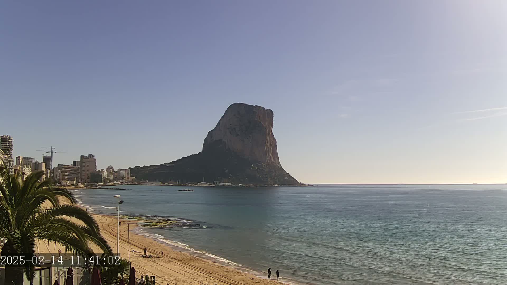 A large, rocky mountain rises from the sea in the distance, visible from a sandy beach with a calm sea under a clear, bright sky.