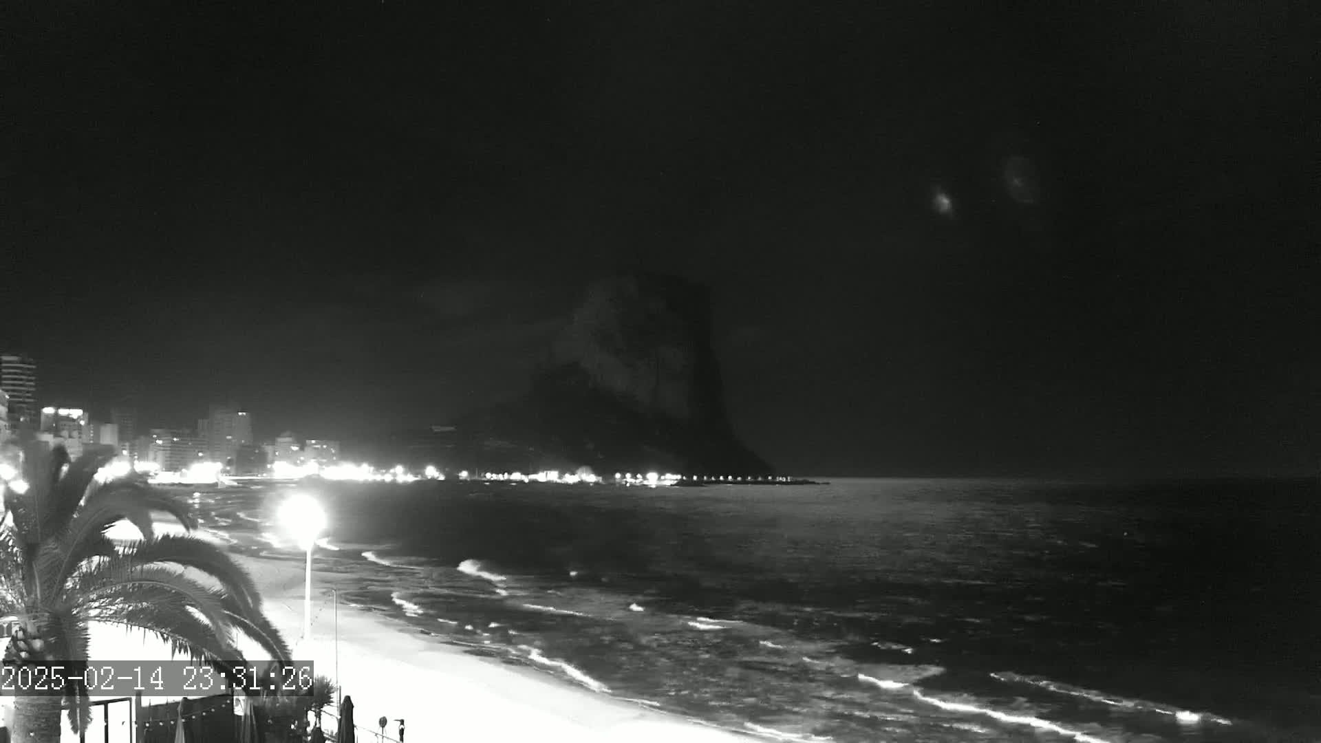 A nighttime, black-and-white view of a beach with gentle waves, a line of illuminated buildings, and a large, dark hill or cliff in the distance under a clear night sky.