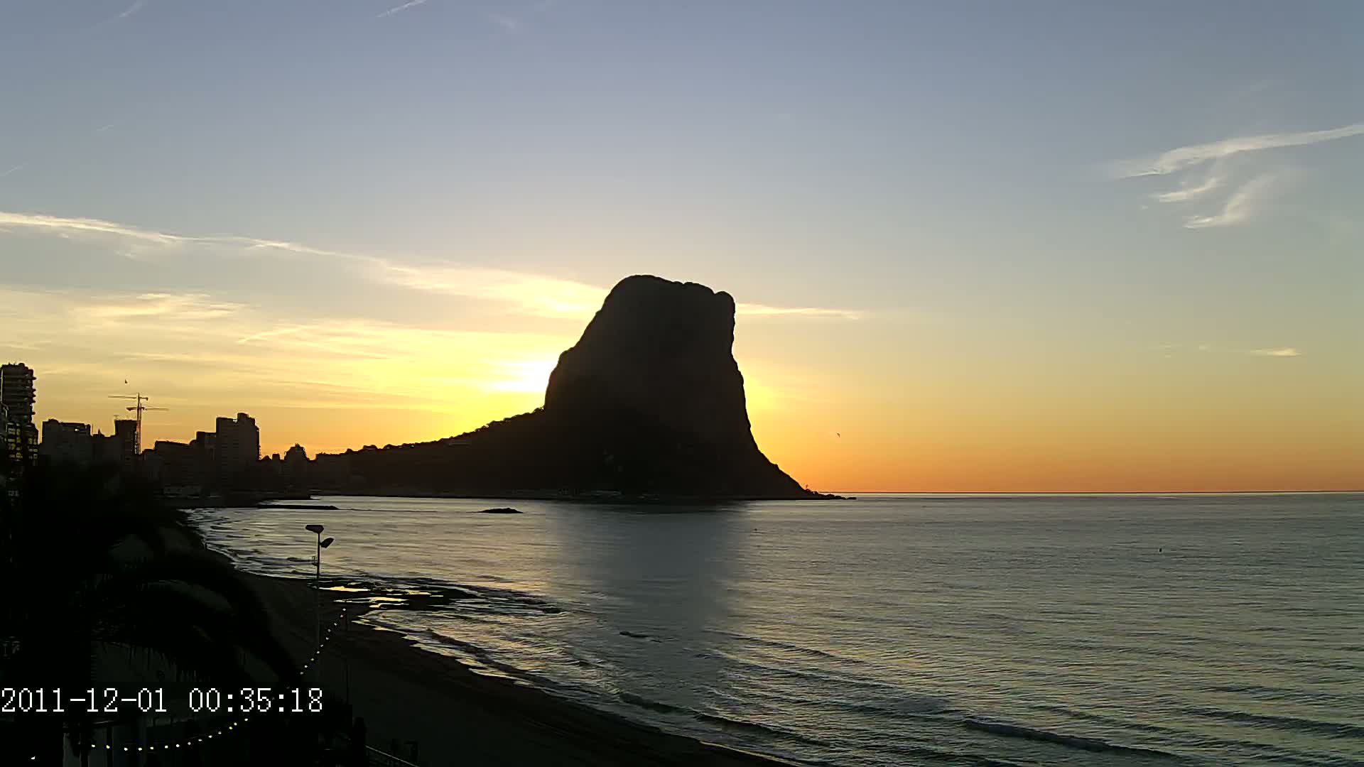 A large, dark silhouette of a rock formation stands prominently against a calm ocean at sunset, with a city skyline visible to the left.