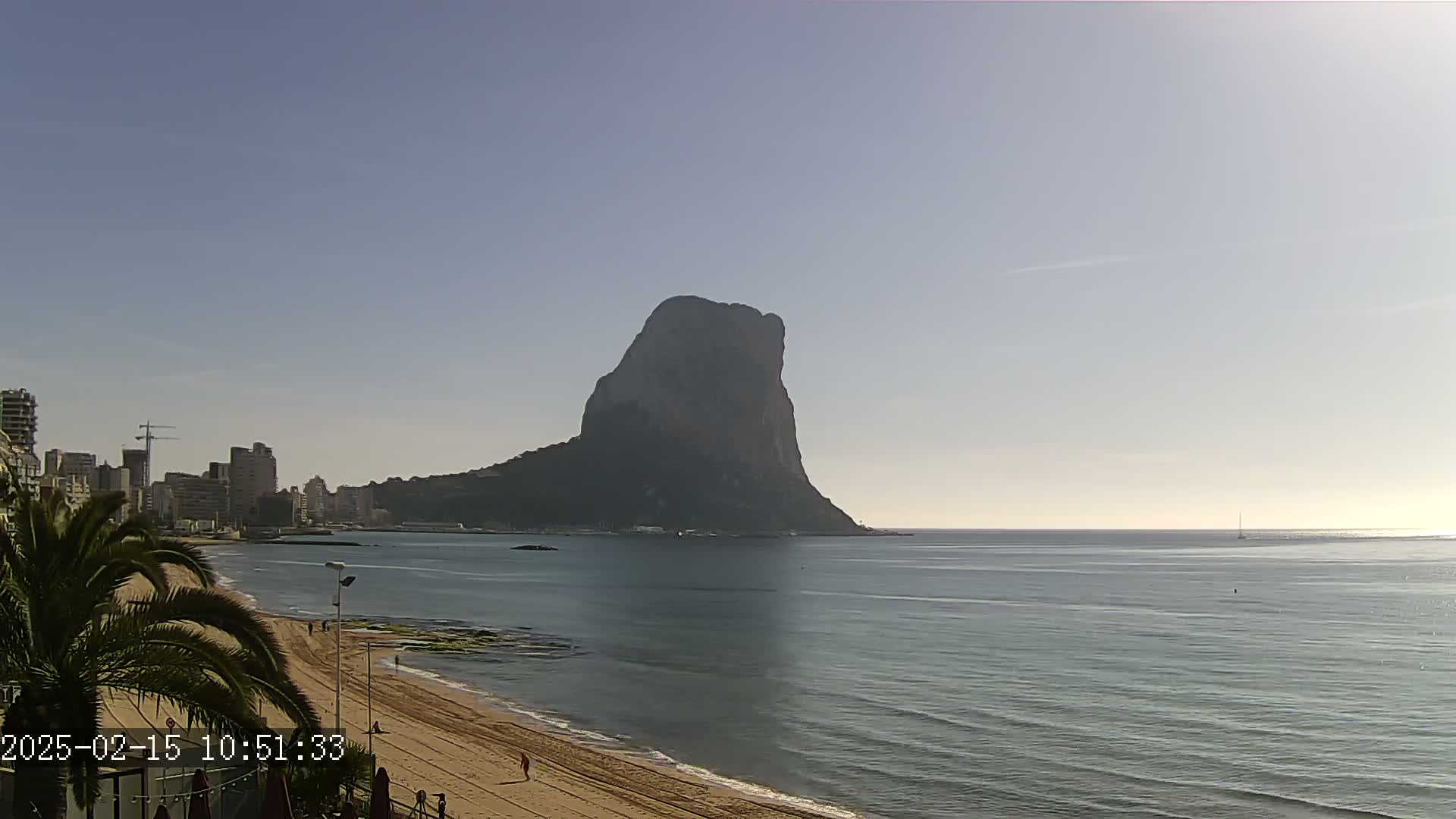 A large, dark rock formation rises from the sea in the distance, with a sandy beach and buildings along a calm, clear coastline under a bright, clear sky.