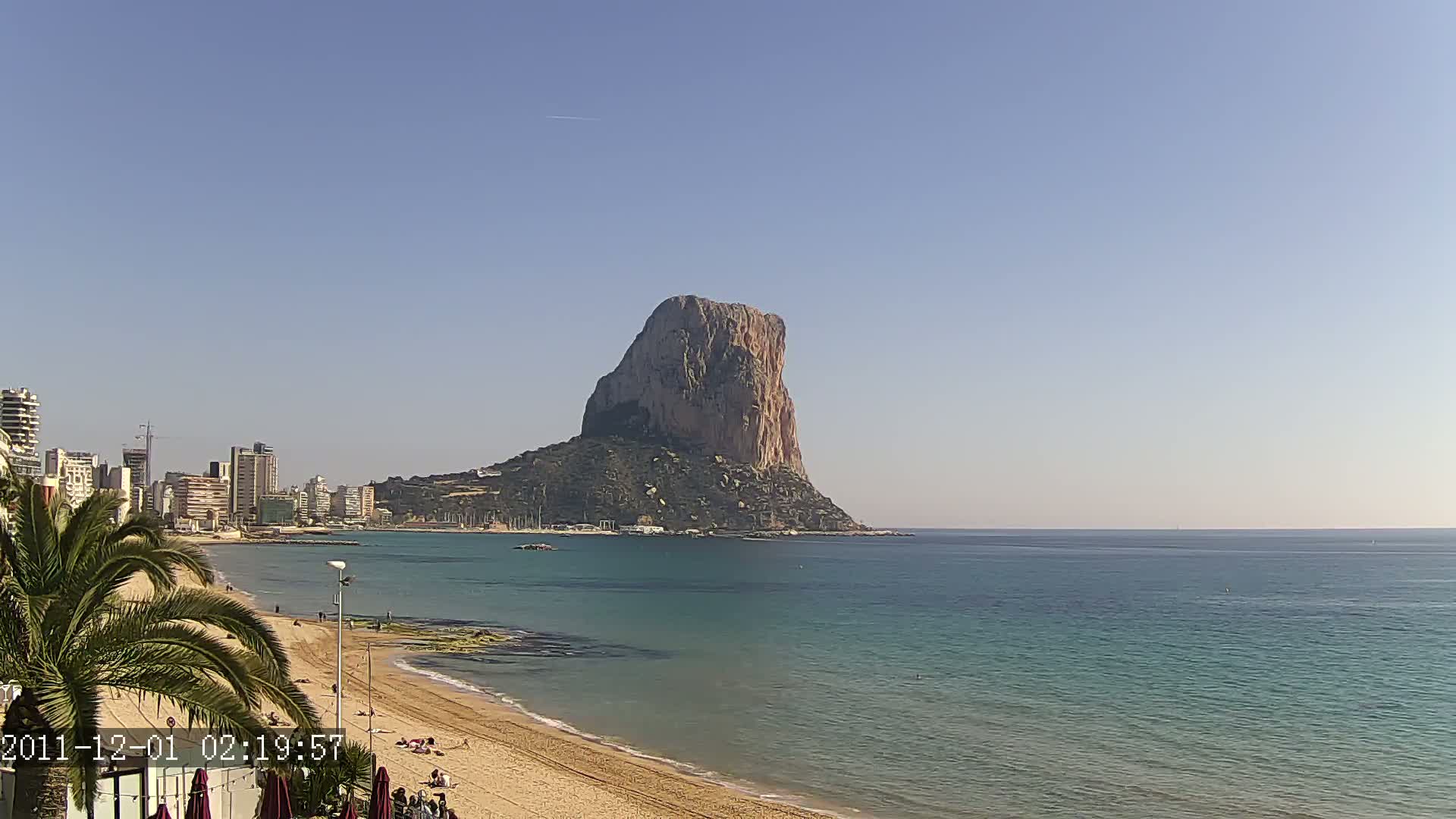 A large, rocky island sits in the calm, blue ocean off a sandy beach, with a city skyline visible in the background under a clear sky.