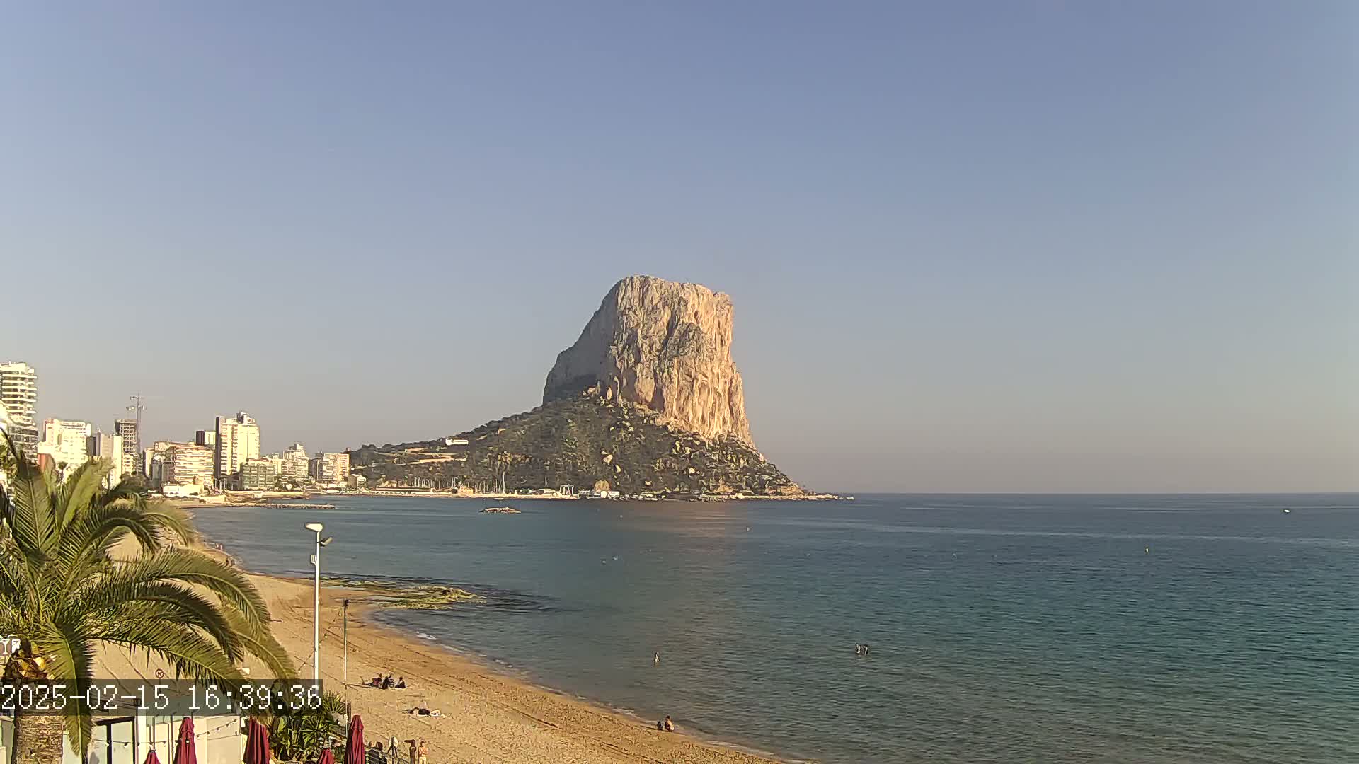 A large, tan rock formation rises from the sea near a sandy beach with a few people on it, under a clear, sunny sky.