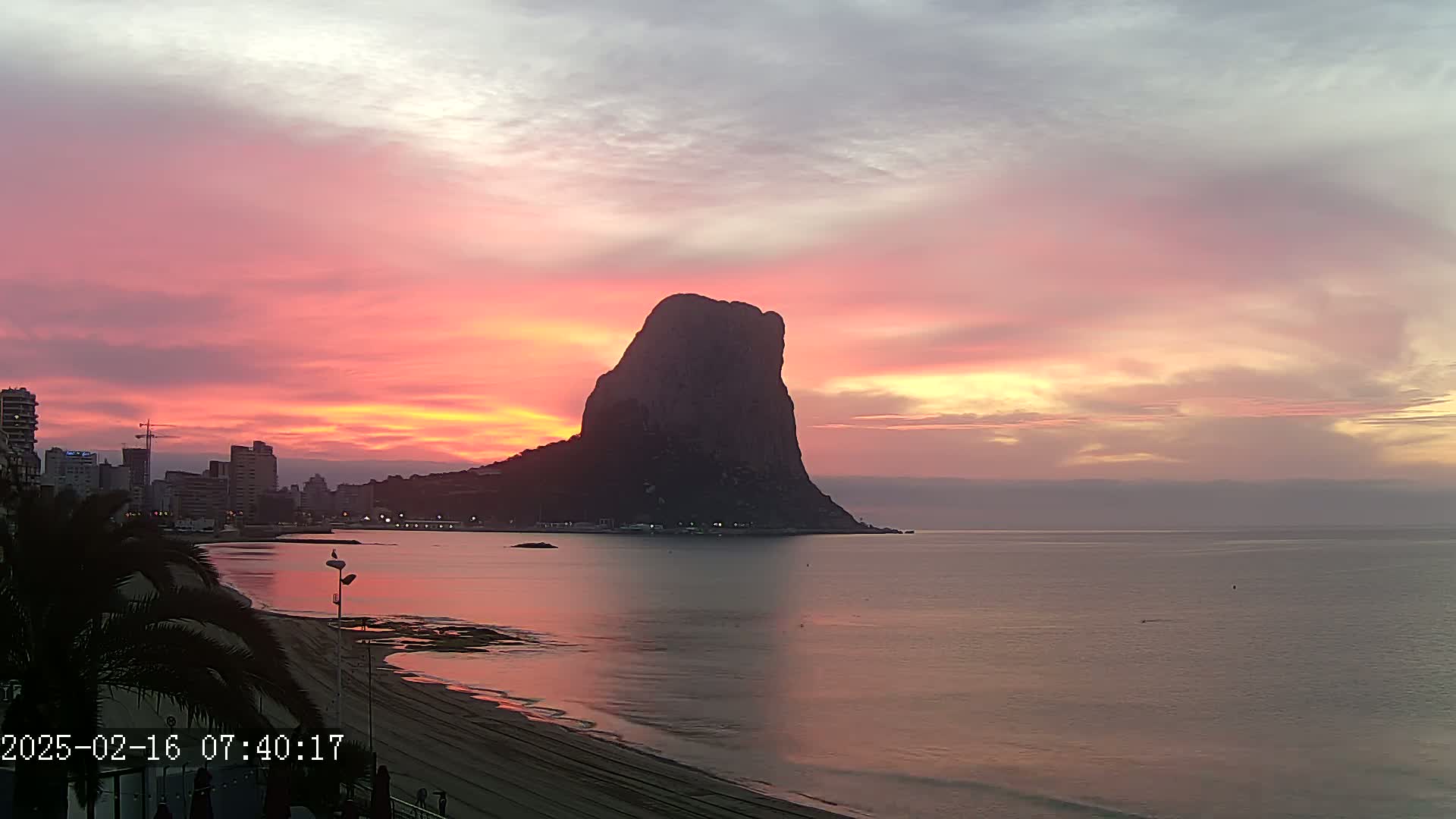 A large, dark rock formation stands prominently against a vibrant pink and orange sunrise over a calm sea, with a city skyline visible in the distance.
