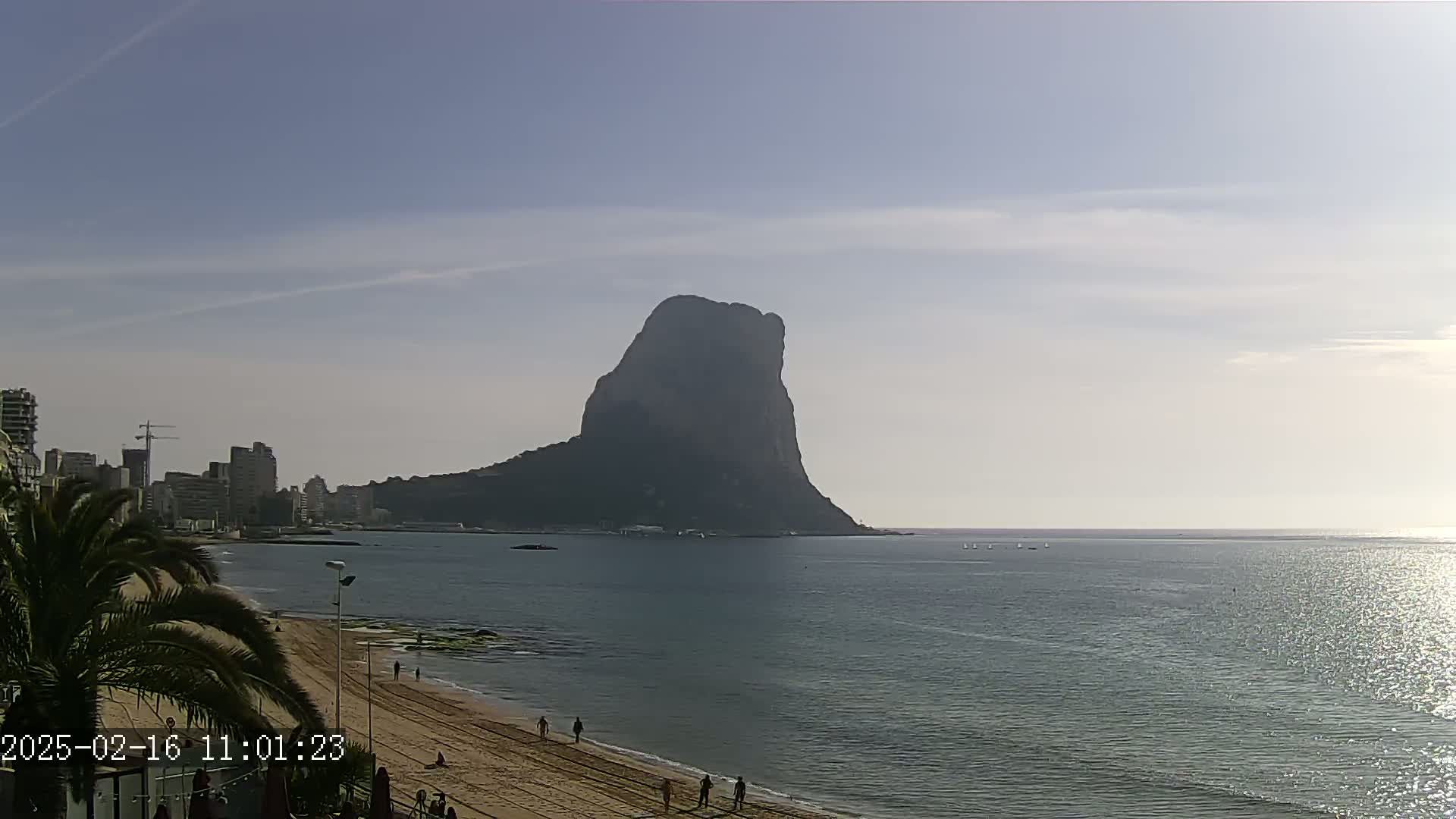 A large, dark rock formation rises from the sea under a mostly clear sky, viewed from a sandy beach with a few people walking and buildings visible in the distance.