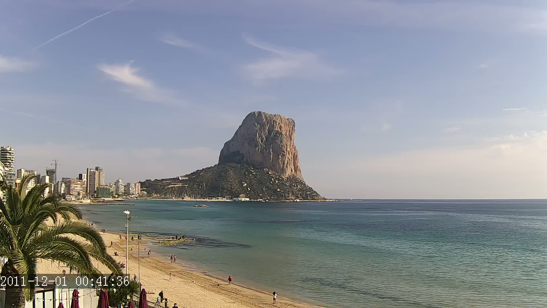 A large, rocky island rises from a calm, teal-colored sea, near a sandy beach with a few people and a cityscape under a mostly clear, sunny sky.