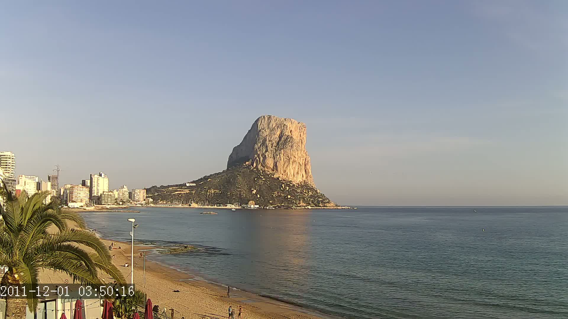 A large, tan rock formation rises from the sea near a sandy beach and a line of buildings under a clear, light-blue sky.