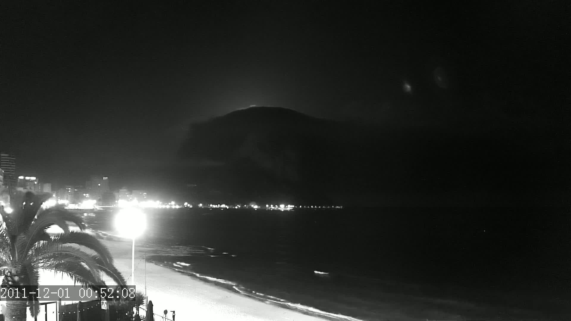 A nighttime, black-and-white view shows a dark mountain in the background, a beach with gentle waves in the foreground, and a city skyline illuminated with lights along the shore.