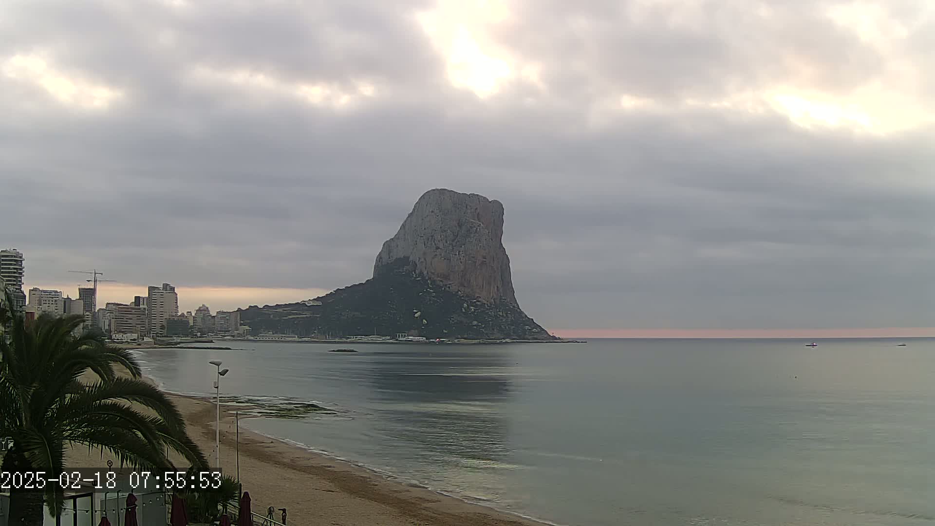 A large, rocky mountain rises from the sea under a cloudy, dawn sky, with a beach and city skyline visible in the foreground.