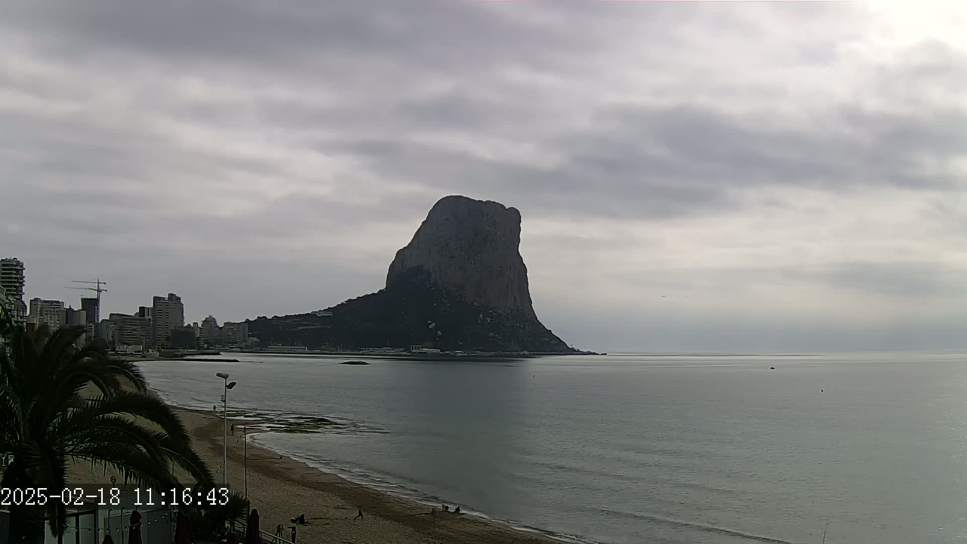 A large, dark rock formation rises from the sea under a cloudy sky, with a beach and buildings visible in the foreground.