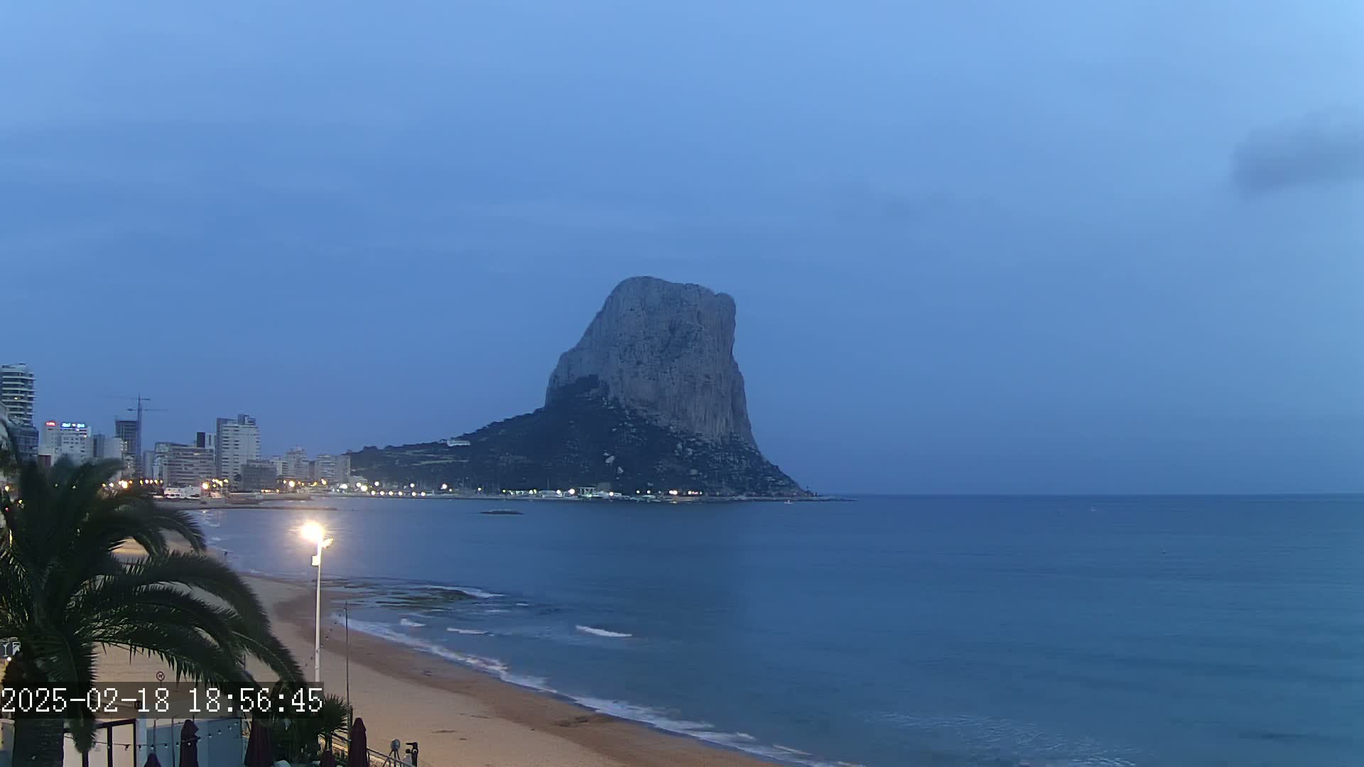 A large, rocky mountain is visible across a calm, blue ocean from a beach at twilight, with a city skyline visible to the left.