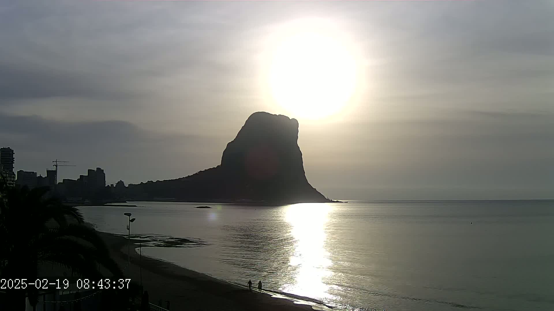 A large, dark, solitary rock rises from the calm sea at sunrise under a mostly cloudy sky, with a city skyline visible in the distance to the left.